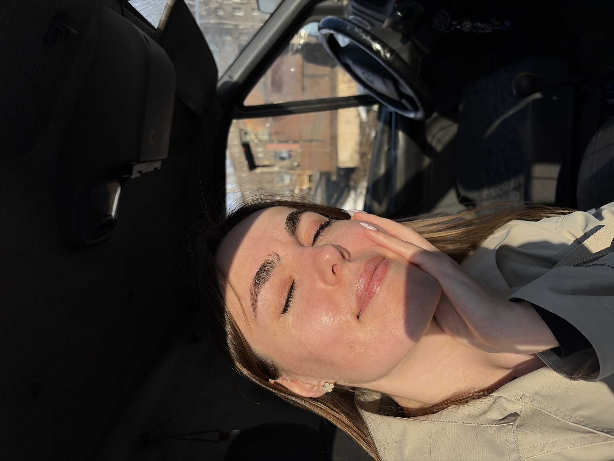 A young woman is reclining in the driver's seat of a vehicle. Her eyes are closed and she is resting her face on her right hand. The vehicle's steering wheel is visible in the upper center of the frame, and the cityscape of Izhevsk, Russia, is seen through the windshield. The woman is wearing a light-colored jacket. The scene is brightly lit.