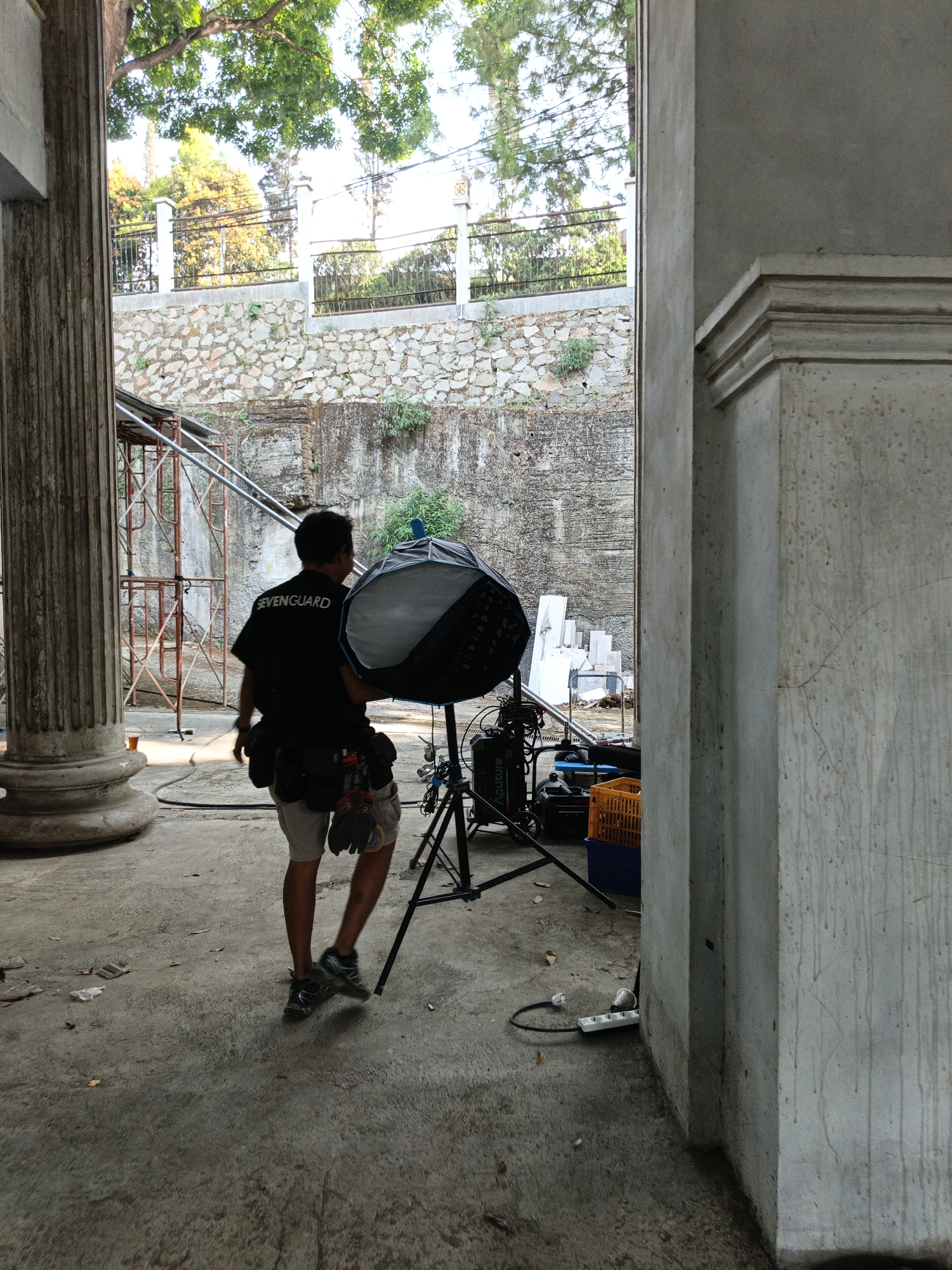 A male photographer, wearing a black t-shirt with "SEVENGUARD" printed on the back, shorts, and athletic shoes, is walking away from the camera. He is carrying a large, hexagonal softbox light on a stand. The softbox is silver and black. He is also wearing a utility belt with pouches and gloves. The scene appears to be an outdoor setting, possibly an abandoned construction site or an old building. There is a large concrete pillar to the left of the frame, and a stone wall in the background. Scaffolding can be seen on the left side of the frame. Electrical equipment, including a generator and extension cords, are present on the ground. A yellow plastic crate and a blue container are visible near the generator. The ground is concrete and appears somewhat worn. The overall atmosphere suggests a work in progress or a candid moment during a photo shoot. The image was taken in Cibogo, Indonesia.