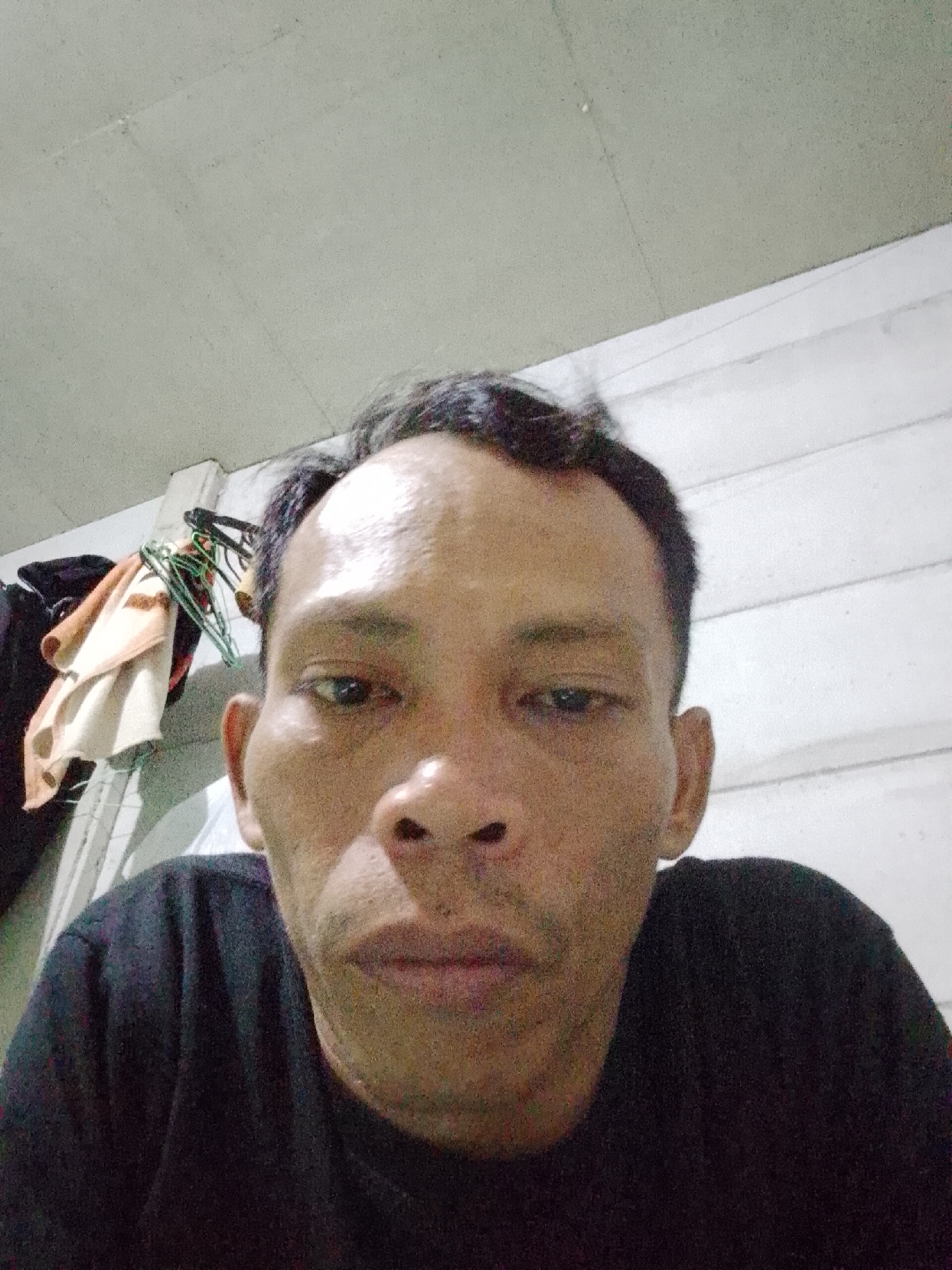 A close-up selfie of a middle-aged Asian man wearing a black t-shirt is displayed. The background features an off-white ceiling and a wall with vertical panels. To the left, clothes hang on hangers. The location is Bukit Ketok, Indonesia.