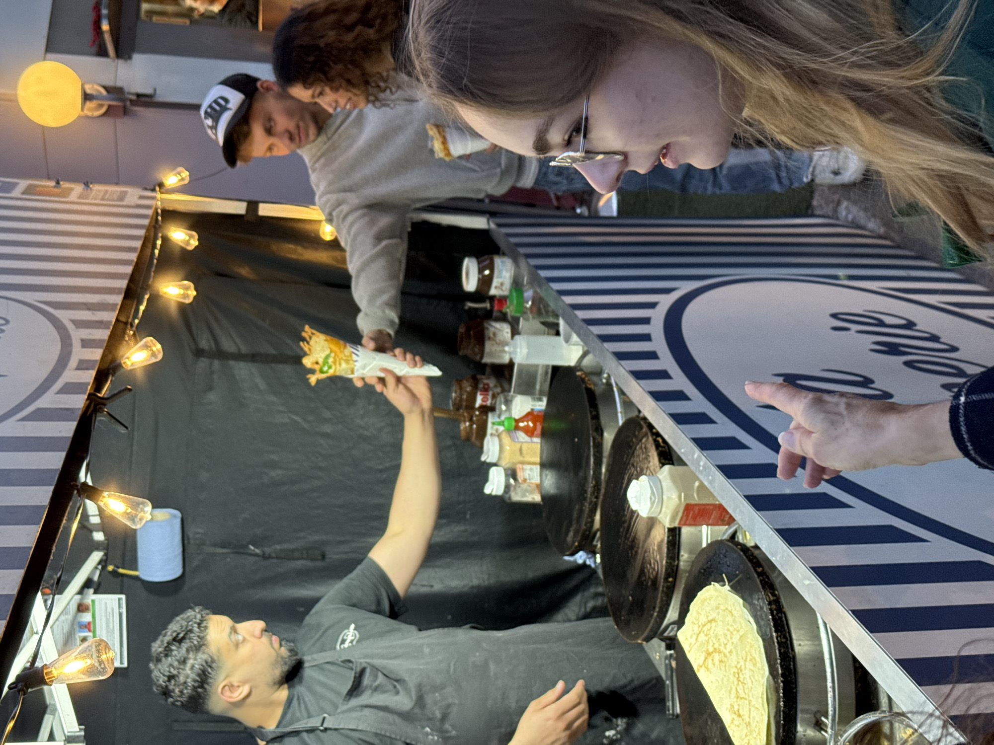 At a food stall in an outdoor setting, a crepe maker presents a filled crepe to customers. The crepe maker, a man with dark hair and beard, wears a dark grey t-shirt and apron. He holds the crepe aloft in his left hand. Customers, including a fair-haired woman with glasses, a man in a baseball cap, and others, observe. A finger points at the food preparation area. The stall is decorated with string lights and contains various jars, bottles, and two crepe cooking surfaces with crepes on them. The stall’s signage features a circular logo with striped patterns. This scene takes place in an unspecified location in the city of London, United Kingdom.