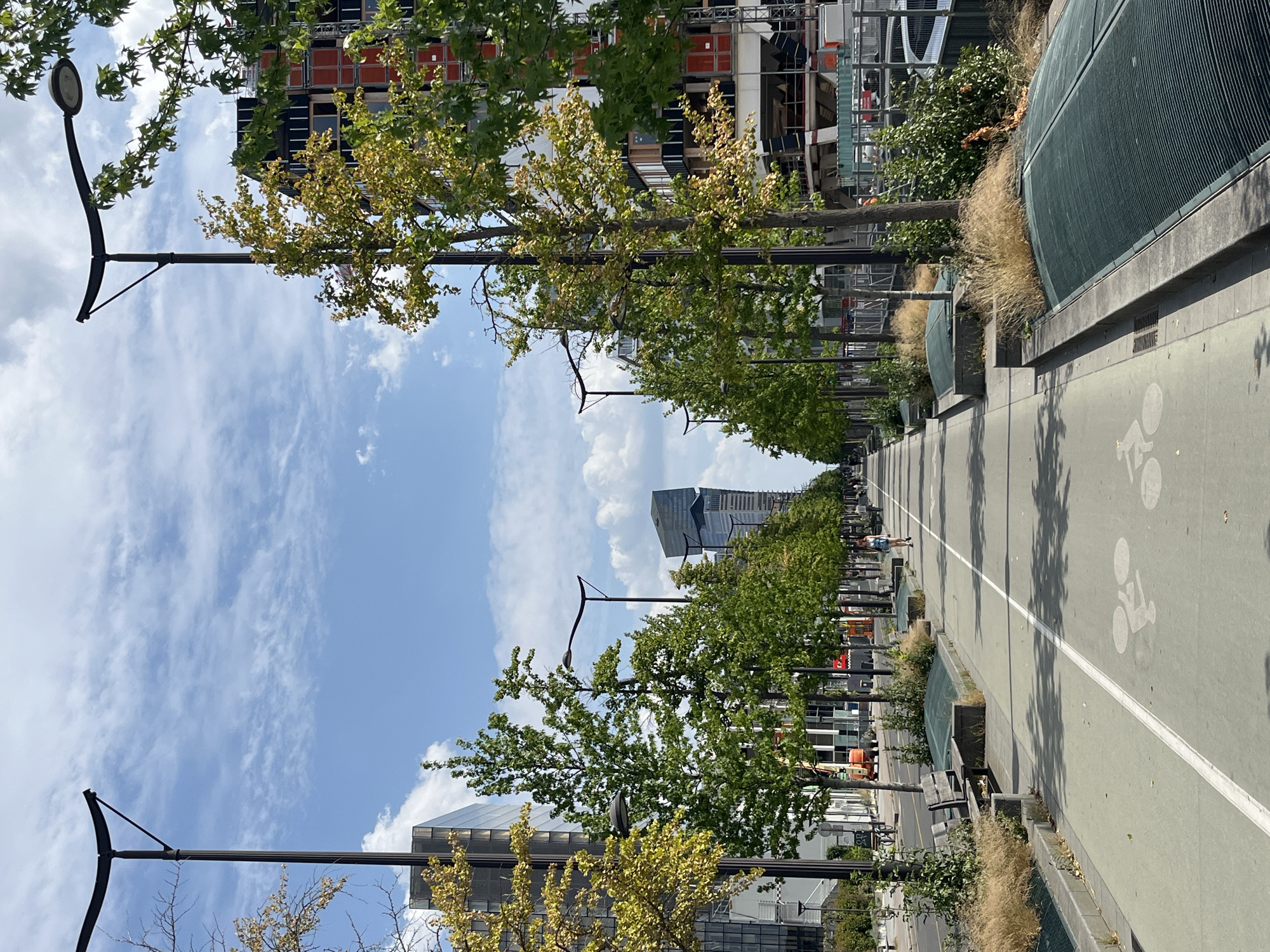 The image depicts a modern urban scene with a distinct focus on a pathway designed for cycling and walking. The pathway is paved and has a clear white line marking its center, with a white bicycle symbol visible on the surface, indicating it is a bike lane. To the right of the pathway is a green, curved structure, possibly a sound barrier or a part of landscaping. Flanking the pathway on the left are rows of trees with green and yellowing leaves, suggesting it might be early autumn. Beyond the trees, modern buildings with glass facades and intricate architectural details are visible. Some buildings appear to be under construction, with scaffolding and cranes in the background.
Several streetlights with a curved design are spaced along the pathway, suggesting an outdoor urban environment. In the distance, a tall, modern skyscraper with a distinctive angular design stands out against a bright blue sky dotted with scattered white clouds. There are a few people visible on the pathway; one person is seen cycling in the distance, and another is walking. The scene suggests a pleasant day, likely daytime, with good weather. The overall impression is one of a well-maintained urban public space designed for recreation and transport. The location is not explicitly identifiable, but the architecture and landscaping suggest a contemporary city.