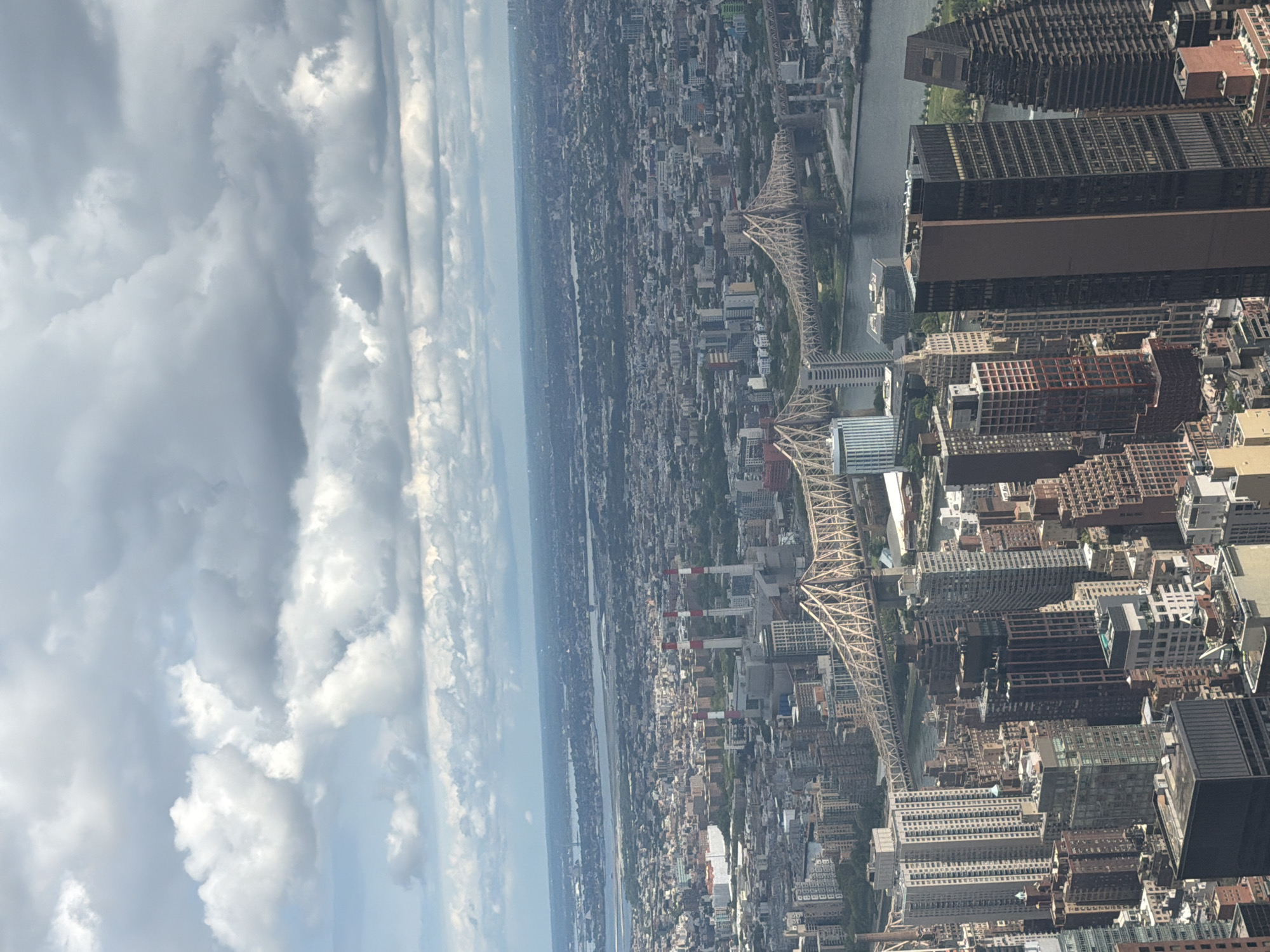 This is an aerial photograph of the New York City skyline, taken from a high vantage point. The image captures a sprawling urban landscape with numerous skyscrapers of varying architectural styles and heights. In the foreground, several tall buildings dominate the view, some with dark facades and others with lighter colors and intricate window patterns.
A prominent feature of the scene is the Queensboro Bridge, a historic cantilever bridge spanning the East River. The bridge's distinctive lattice-work structure is clearly visible as it connects Manhattan to Queens. The East River itself is visible, with dark water reflecting the overcast sky.
Further into the distance, the cityscape extends to the horizon, with a vast expanse of buildings and smaller structures. The distant landscape appears hazy due to atmospheric conditions. In the mid-ground, a power plant with three tall smokestacks is visible, adding an industrial element to the urban panorama.
The sky is filled with dramatic clouds, ranging from bright white cumulus formations to darker, more diffused gray clouds, suggesting a partly cloudy or overcast day. The lighting conditions are somewhat muted due to the cloud cover, but the city is still well-illuminated. The overall impression is of a dense, dynamic urban environment under a significant sky.