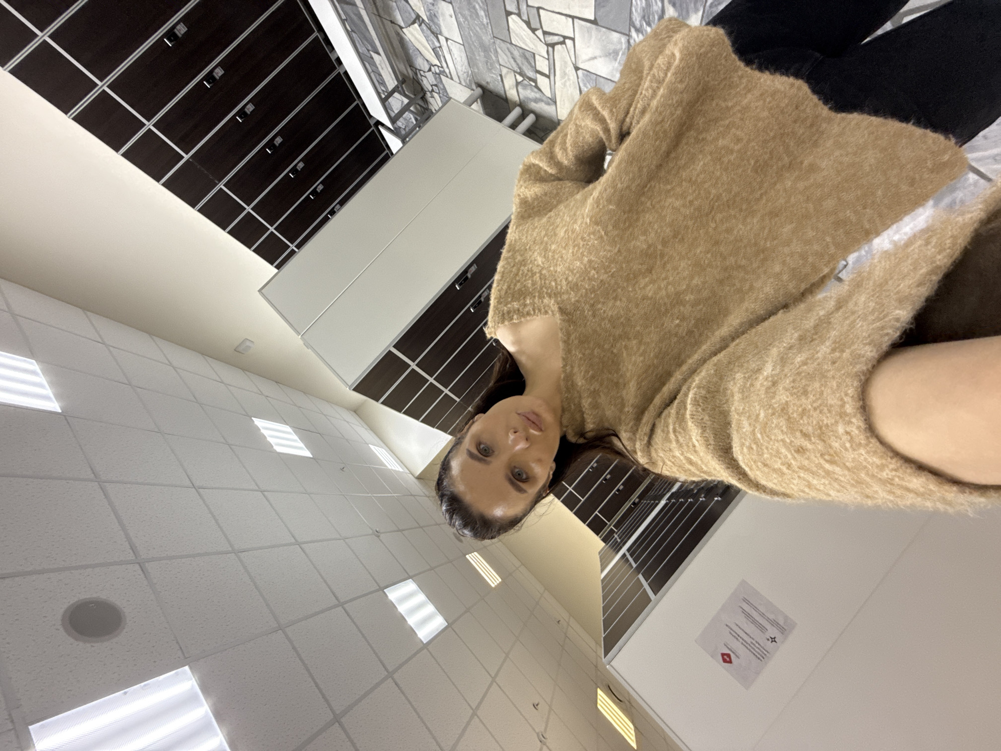 A woman in a brown sweater and black pants is the primary subject of the photograph. She appears to be inside a building. The environment includes a tiled ceiling with recessed lighting, dark brown lockers with silver trim, and white cabinetry. A sign with red and black symbols is visible on a white wall. The location is potentially Chaikovsky, Russia.