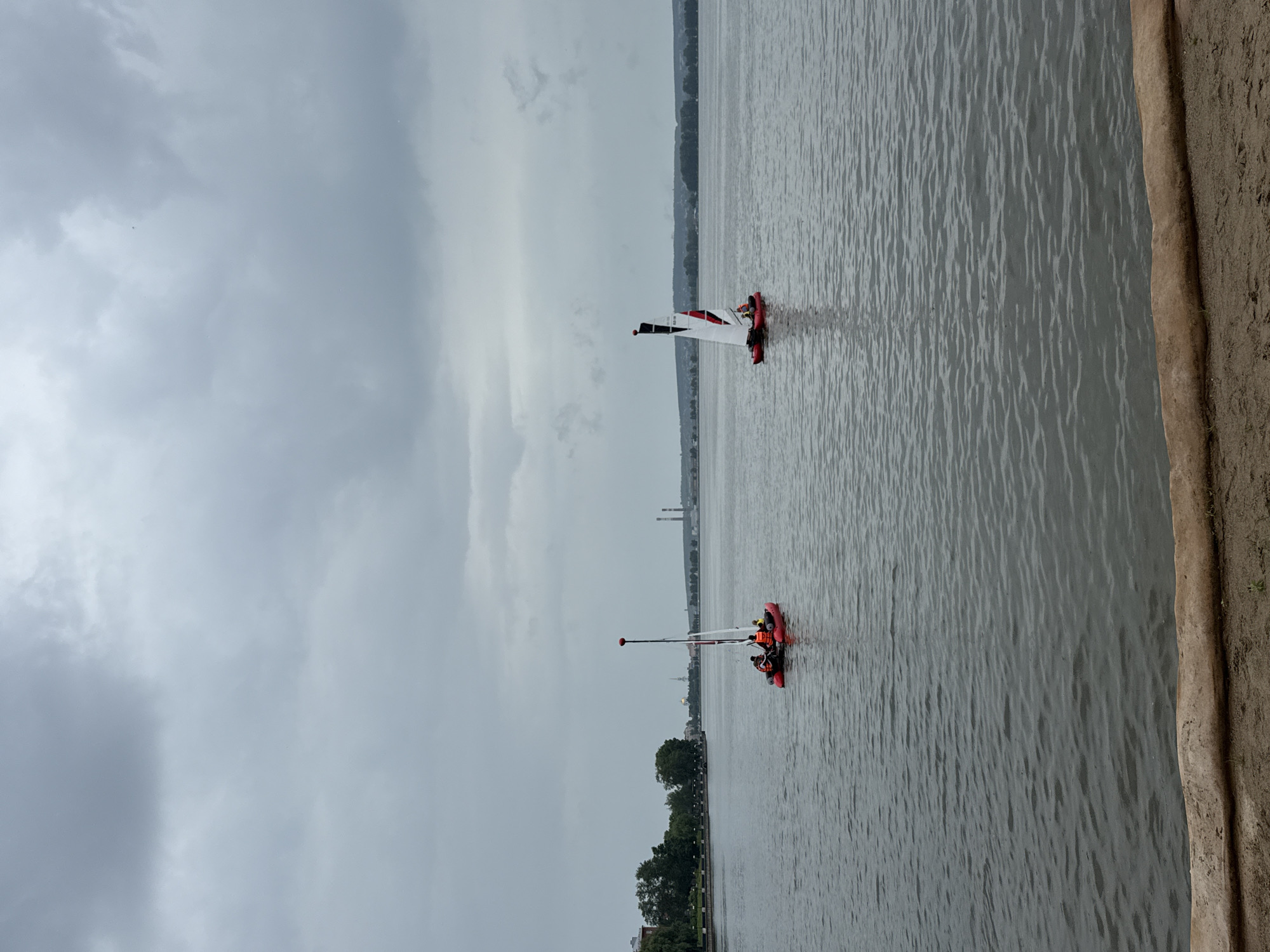 This image depicts a cloudy day on the Votkinsk Reservoir in Votkinsk, Russia. Two small sailboats are on the water. The sailboat on the left is red and has three people on board, all wearing life jackets. The sailboat on the right is also red and has one person visible. The background shows a treeline, some buildings, and industrial smokestacks. The foreground is a sandy bank with a log.