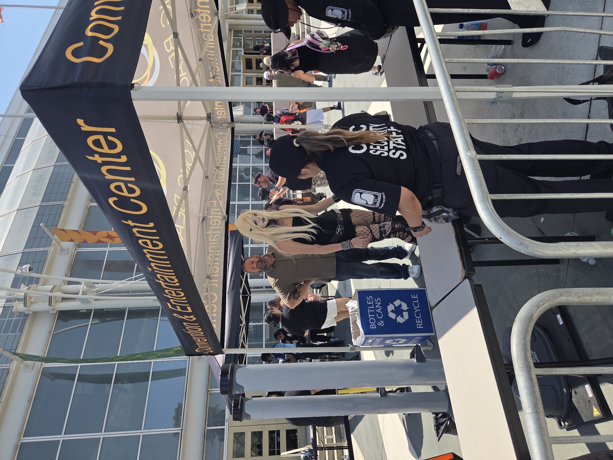 In Long Beach, California, outside a convention center, a security checkpoint is set up. Two security personnel, one in the foreground wearing a black shirt with "CSC SECURITY STAFF" and a black hat, and another to her right in a black shirt and hat, monitor the entrance. In front of them, a woman with pale makeup and blonde hair, dressed in gothic attire including fishnet stockings, stands near a man in a brown shirt and jeans. A blue recycling bin labeled "BOTTLES & CANS RECYCLE" is positioned between the security and the attendees. Two tall, gray cylindrical metal detectors stand to the left. The environment suggests an outdoor event or convention, with a large building featuring glass windows in the background, under a clear sky.
