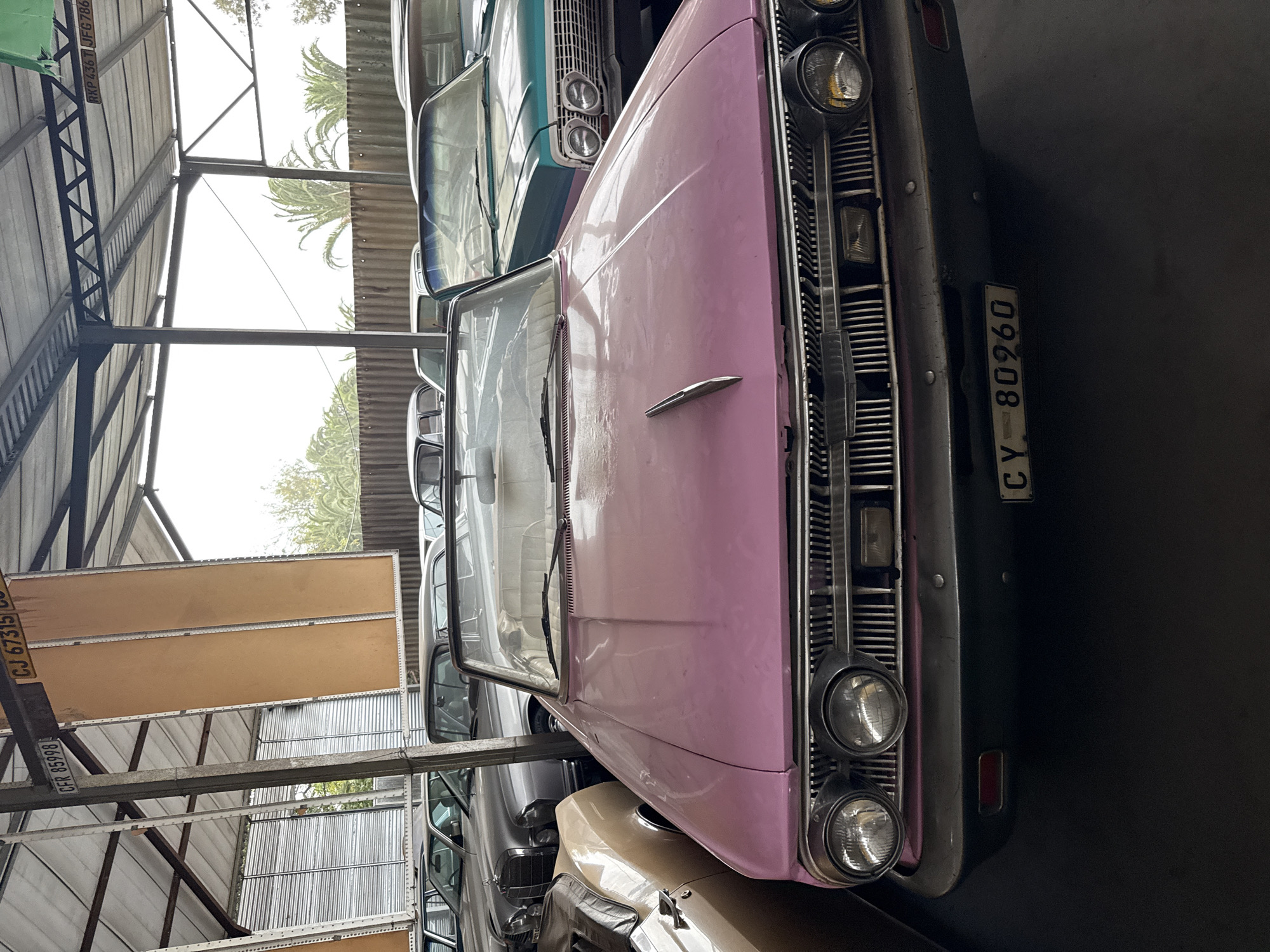 The image shows a collection of vintage cars stored indoors, possibly in a warehouse or garage. The cars are parked close together, with the front of a pink car dominating the foreground. The pink car has a visible license plate that reads "CY 80960". The grills, headlights, and hood of the car are clearly visible. Behind the pink car, other vintage cars in various colors (such as turquoise and silver) are partially visible. The location is suggested to be Kraaifontein, South Africa. The building appears to be a storage facility with corrugated metal walls and ceiling beams. There is a structure of metal beams and supports visible in the lower left. The environment is dimly lit.