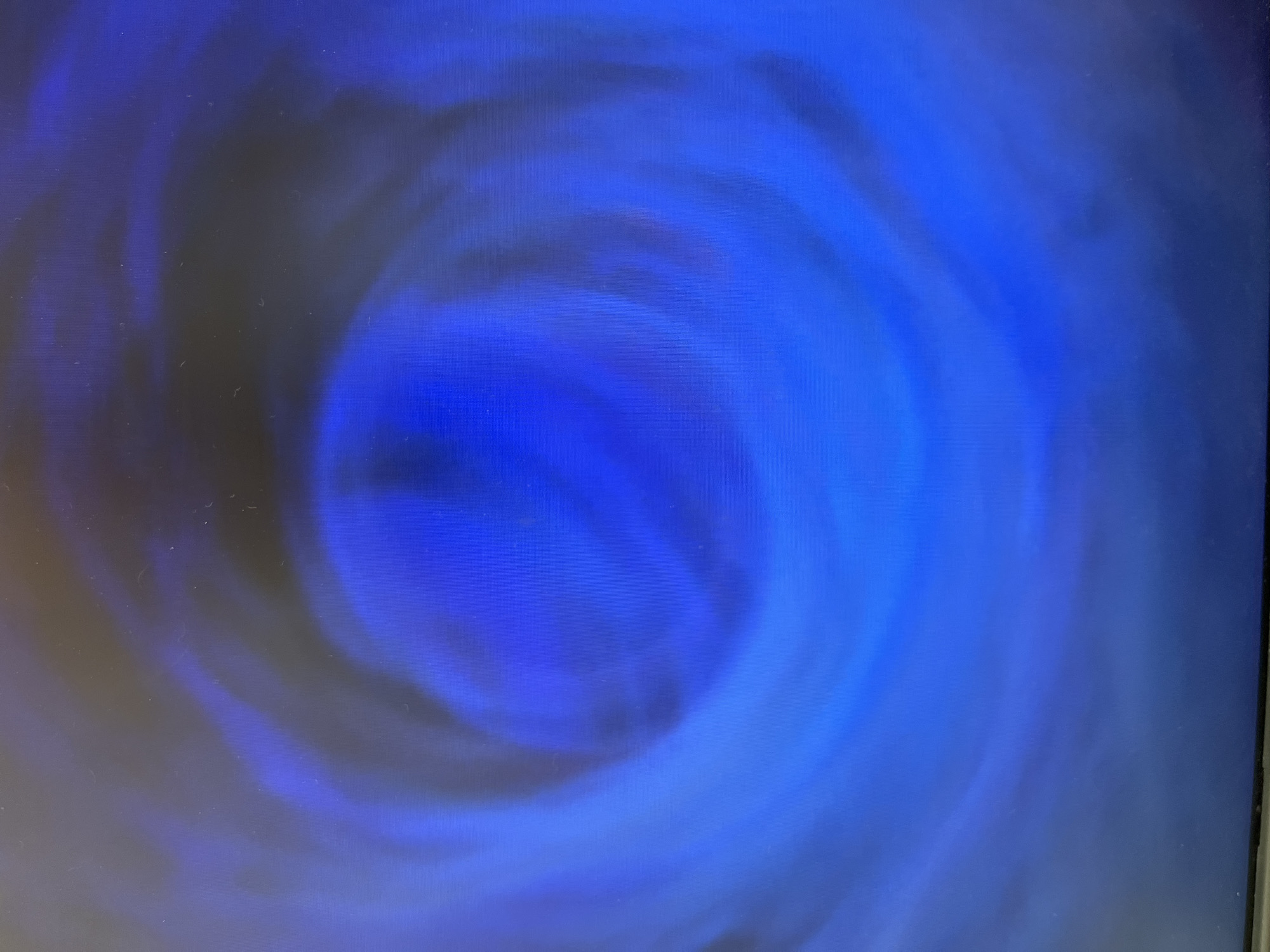 The image shows a swirling vortex of blue and dark gray hues. The pattern is circular, with the darkest area concentrated in the center and the blue shades radiating outwards in a spiral. No specific location, people, subjects, objects, actions, interactions, or environmental details are discernible.