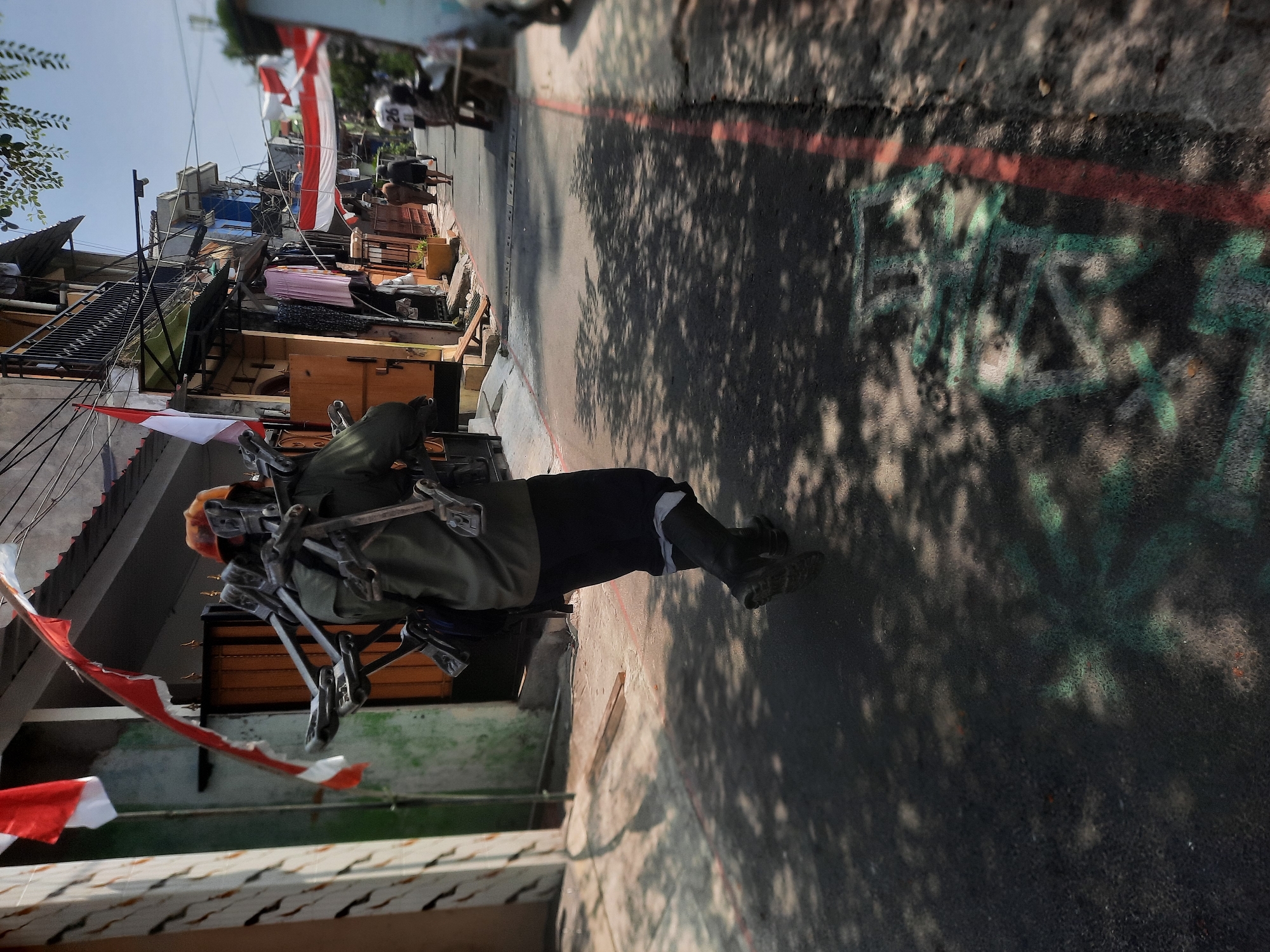 In a narrow, sun-dappled alleyway in North Jakarta, Indonesia, a person is walking away from the camera, carrying a large, metal contraption strapped to their back. The contraption appears to be made of several interconnected metal pieces, possibly a frame or a rack, with some clamps or holders attached. The individual is wearing a green jacket, dark pants, and black boots. On their head, they have an orange-colored cap.
The alley is lined with tightly packed, multi-story buildings. Several Indonesian flags, red and white, are displayed prominently on the buildings, some hanging from balconies and others attached to walls, suggesting a patriotic or celebratory occasion. Laundry is visible hanging from clotheslines and balconies. The ground of the alley is asphalt with some red and white painted lines, and there is graffiti visible in green on the surface in the foreground.
In the background, other people are visible walking further down the alley, and a scooter or motorcycle is parked on the side. The overall impression is of a densely populated urban environment, with signs of daily life and national pride. The sunlight casts dappled shadows of tree leaves across the ground, adding a sense of depth and texture to the scene.