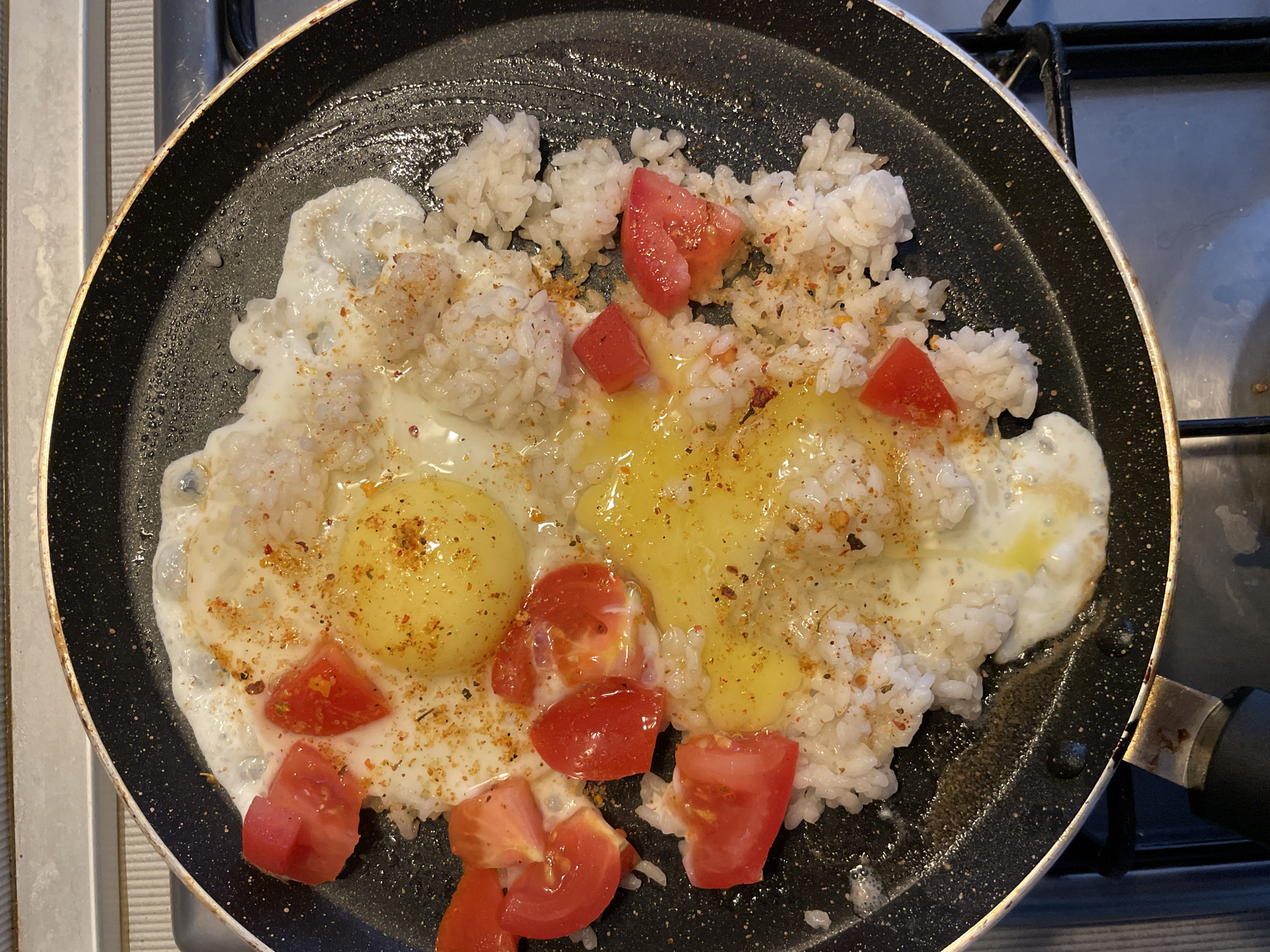 A top-down, close-up shot captures a black non-stick frying pan on a gas stovetop. The pan contains a dish of rice, two fried eggs with runny yolks, and chopped tomatoes. The dish is seasoned with what appears to be red pepper flakes and other spices. The background shows a portion of a stainless steel stovetop and part of a black burner grate. The scene is an indoor kitchen environment. The exact location within the city of Sochi, Russia, cannot be determined from the image alone.