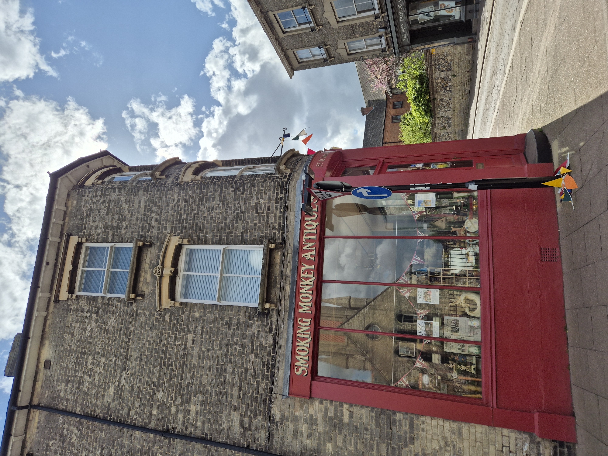The image shows the exterior of "Smoking Monkey Antiques" shop, located in West Suffolk, United Kingdom. The shop, painted red, has large windows displaying various items for sale and reflects the image of a church building across the street. The shop's name is painted vertically on the window frame. A brick building is attached to the left of the shop, featuring two windows. Above the shop, a street sign and colorful flags are visible. The sidewalk is adjacent to the shop on the right. The sky above features scattered clouds.