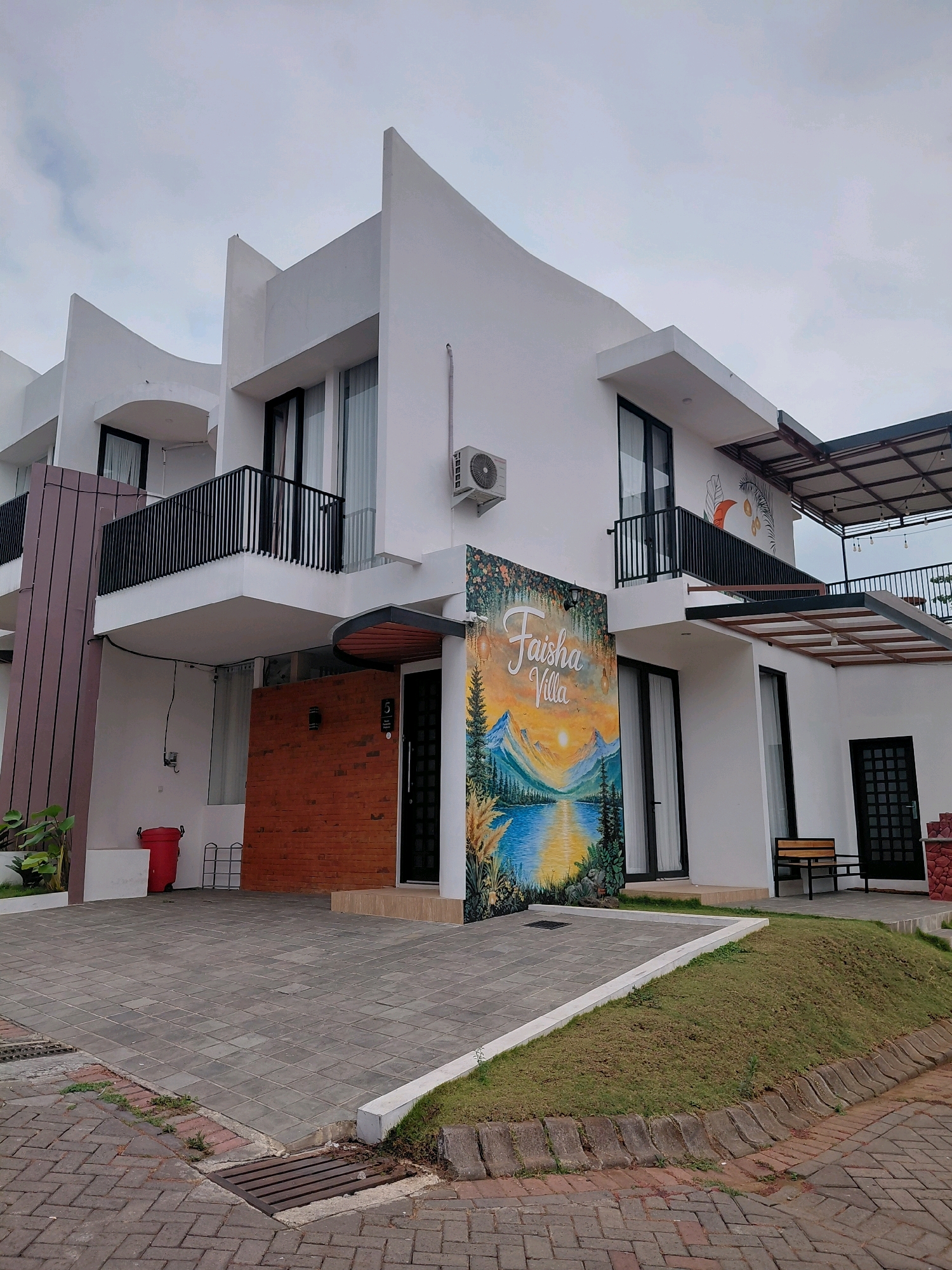The image depicts the exterior of "Faisha Villa" in Batu, Indonesia. The villa is a modern, two-story building with a white facade, characterized by sharp geometric angles and balconies with black railings. A large mural featuring a serene landscape with mountains, a lake, and a sunset adorns one side of the villa's exterior, with the text "Faisha Villa" prominently displayed within the artwork. The building is surrounded by a paved driveway made of interlocking grey bricks and a small, grassy slope. A red trash bin and a metal rack are visible near the entrance on the left side, while a wooden bench is placed outside on the right. The sky is overcast, suggesting cloudy weather. No people are visible in the scene.
