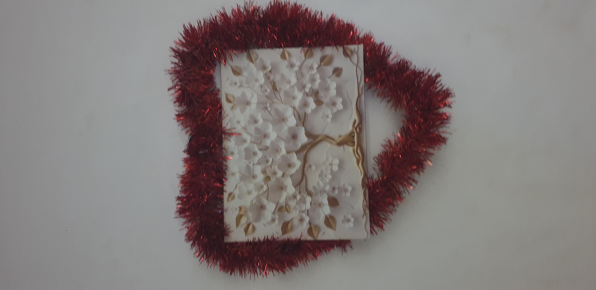 A rectangular, three-dimensional decorative plaque featuring a stylized tree with a brown trunk, exposed roots, numerous white blossoms, and scattered golden leaves is affixed to a plain, light grey wall. The plaque is centrally bordered by a heart-shaped arrangement of shimmering red tinsel garland. The scene is illuminated evenly, suggesting an indoor setting. The specific location within Narayi, Nigeria, is not discernible from the image.