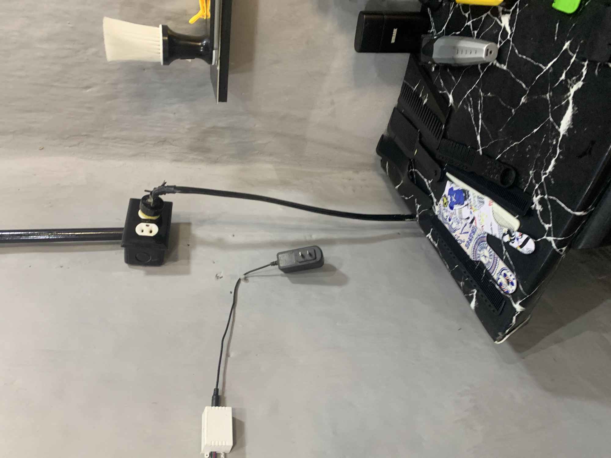 This image captures an indoor scene, likely a utility area or workshop, with a focus on electrical connections and various objects mounted or placed against a light gray wall.
In the mid-left, a black electrical outlet box with a single white duplex receptacle is surface-mounted on the wall. A black conduit or cable runs horizontally into this box. A black power cord is plugged into the upper socket of this outlet, but the insulation around the plug's connection to the cord appears damaged or frayed, exposing wires. This cord extends to the right, connecting to a larger black object.
On the right side of the frame, angled towards the viewer, is a flat, rectangular object with a distinct black and white marble-like pattern. This object, possibly a laptop or a decorative panel, has several black combs of different sizes placed on its surface. Also on this object is a silver electric shaver or hair trimmer, with the brand "WAHL" faintly visible on its side. Below these items, colorful stickers or decorative papers are visible on the marble-patterned surface.
Further down and to the left of the marble-patterned object, a black power adapter, often referred to as a "wall wart," hangs by its cable. It is connected to a small, white rectangular electronic device, which has multiple colorful wires extending from its bottom.
In the upper left corner, a white-bristled brush with a black handle is mounted on a black surface against the wall. The general lighting suggests an indoor setting. The visible damaged wiring at the outlet presents a potential safety hazard. The combination of electrical components and grooming tools suggests a functional space, possibly a home workshop or a barber/salon setup. The location context for this image is La Tagua, Venezuela.