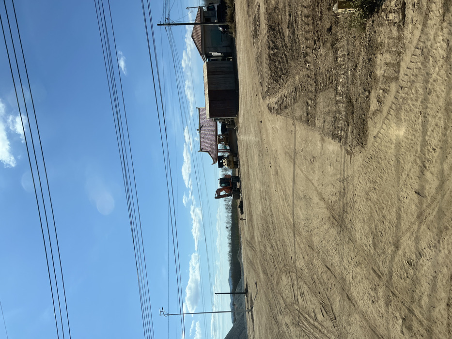 A sunny scene captures a dirt road with visible tire tracks. Adjacent to the road, on the left, stands a building with a distinctive red-tiled roof. Construction vehicles, including an excavator, are parked nearby. High-voltage power lines stretch across a blue sky dotted with clouds. In the distance, a line of trees and a gravel pile are visible. The foreground is dominated by the textured surface of the dirt road, showing signs of heavy use.