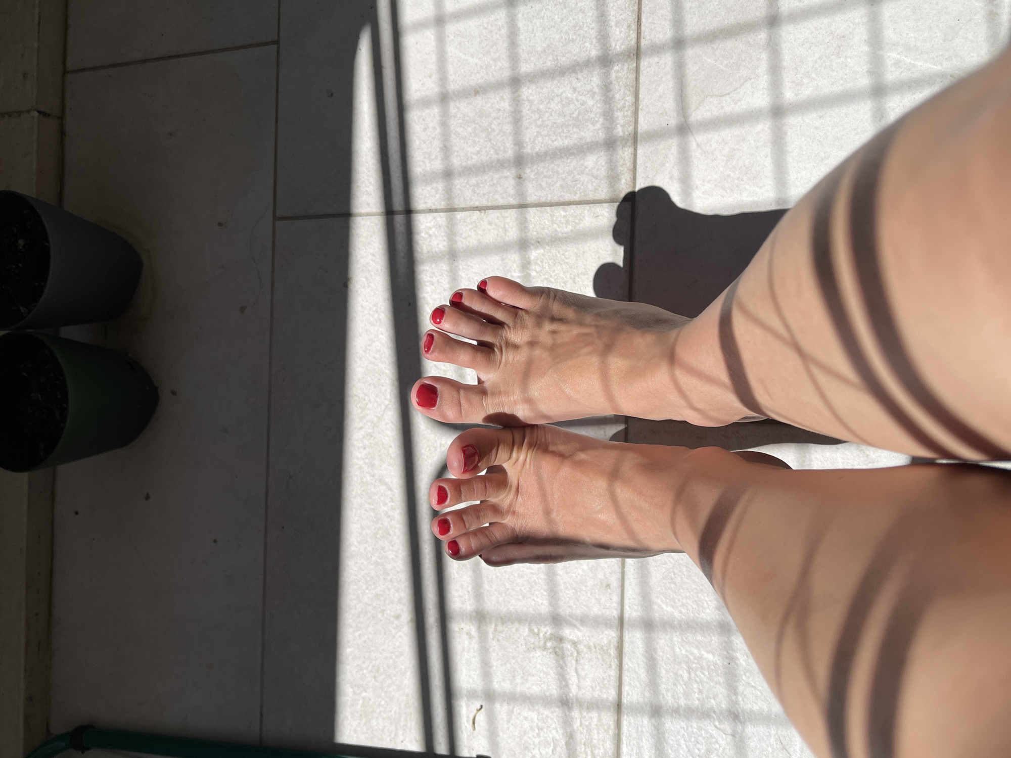 The image shows a pair of feet, with red nail polish on the toenails, resting on a tiled surface. The lighting suggests an outdoor environment, with shadows cast from what appears to be a fence or railing. Two potted plants are visible in the background. The feet are positioned in the center of the frame, with the legs extending downwards out of view. The overall impression is one of relaxation and a sunny day. The scene is devoid of any specific landmarks to indicate its location in the city of Unknown.