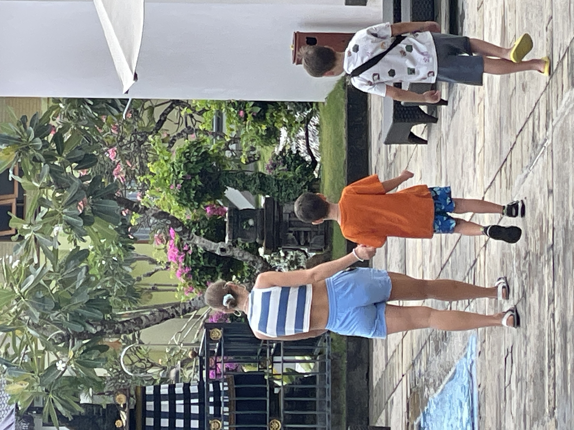 In Kuta Selatan, Indonesia, a woman in a blue and white striped crop top and light blue shorts is walking away from the camera, holding hands with a young boy in an orange t-shirt and blue patterned shorts. Beside them, another young boy in a white t-shirt with a pattern of small colorful shapes and grey shorts walks in the same direction, carrying a black strap across his chest. They are on a paved walkway bordered by lush greenery and trees with pink flowers. In the background, there is a black and white checkered structure, a metal fence, and part of a white building with a partially unfurled umbrella. A small red trash can is visible to the right of the frame. The scene appears to be an outdoor area, possibly a resort or a courtyard.