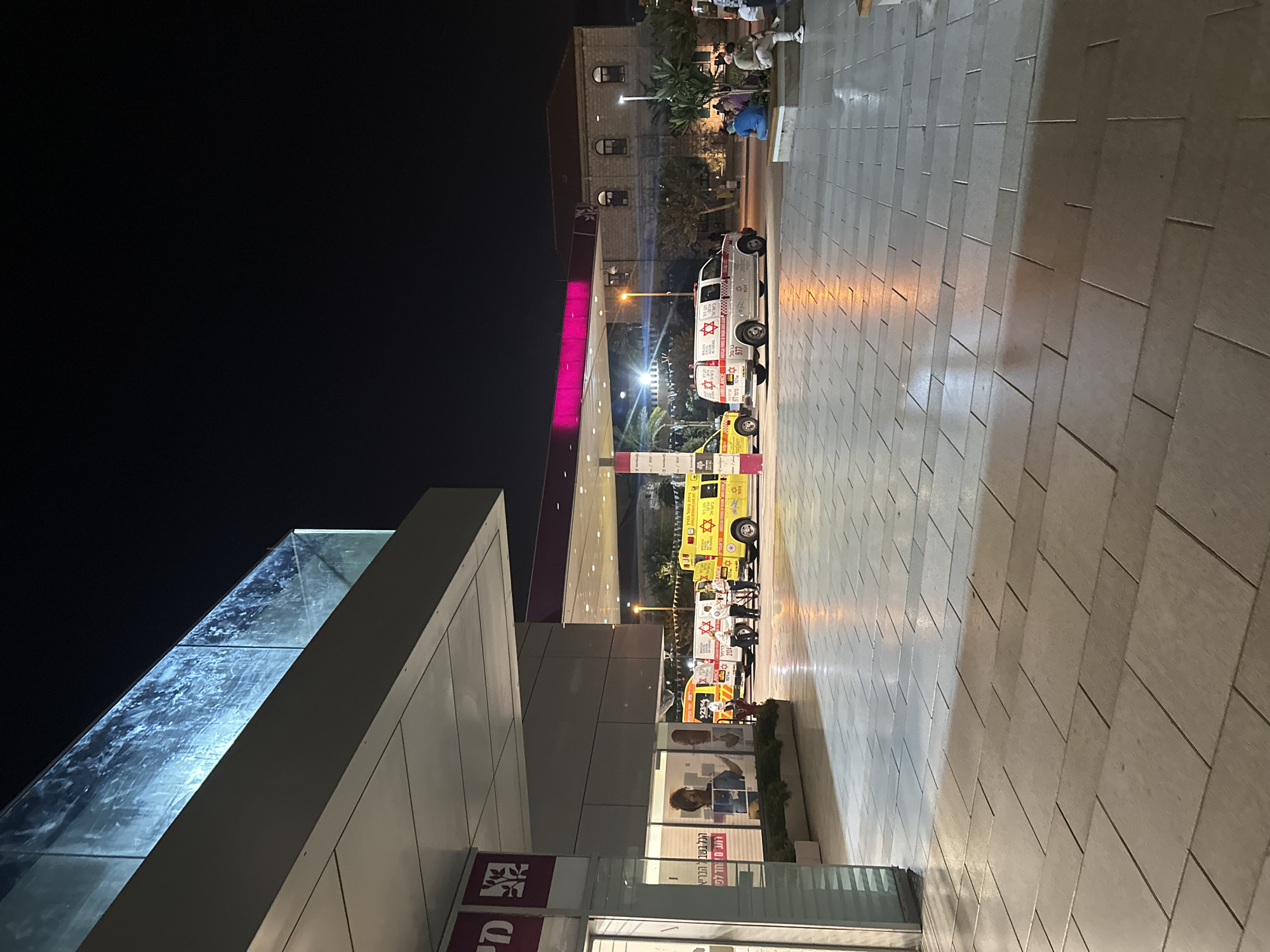 The image depicts an outdoor scene at night, likely in Haifa, Israel, with several ambulances parked under a canopy. The ambulances are yellow and white, bearing the Star of David and text that appears to be in Hebrew. A modern building with a glass balcony overhang is visible on the left, with some signage in Hebrew. In the background, a more traditional stone building with arched windows is illuminated. Several people are seen near the ambulances and sitting on a low wall to the right. The ground is paved with large, rectangular tiles, some of which are wet and reflecting the artificial lights. The overall atmosphere is one of a nighttime operation or event involving emergency services.