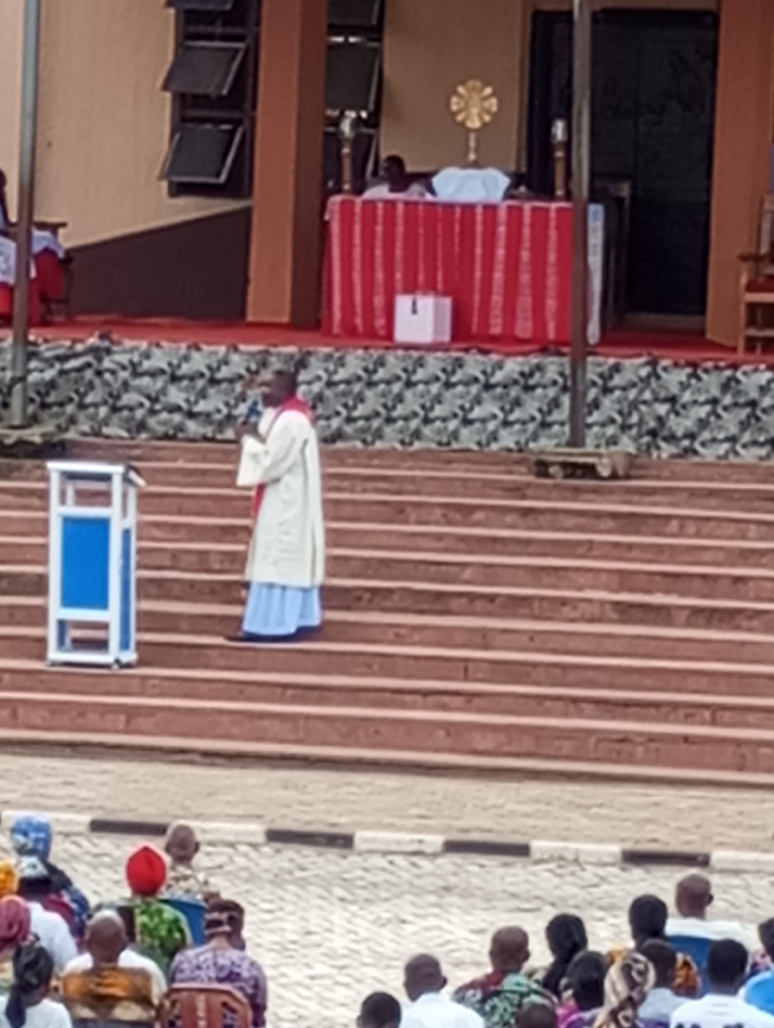 The image depicts a man, likely a priest, speaking from a podium on a staircase in front of a building. The man is wearing a white robe over a light blue garment, with a red sash. A blue and white podium is to his left. In the background, on an elevated platform, there is a table covered with a red and white cloth, on which sits a gold cross or religious symbol. In front of the staircase is a seated audience, presumably listening to the speaker. The location is identified as Onitsha, Nigeria, due to the architectural style of the building and the attire of the people present.
