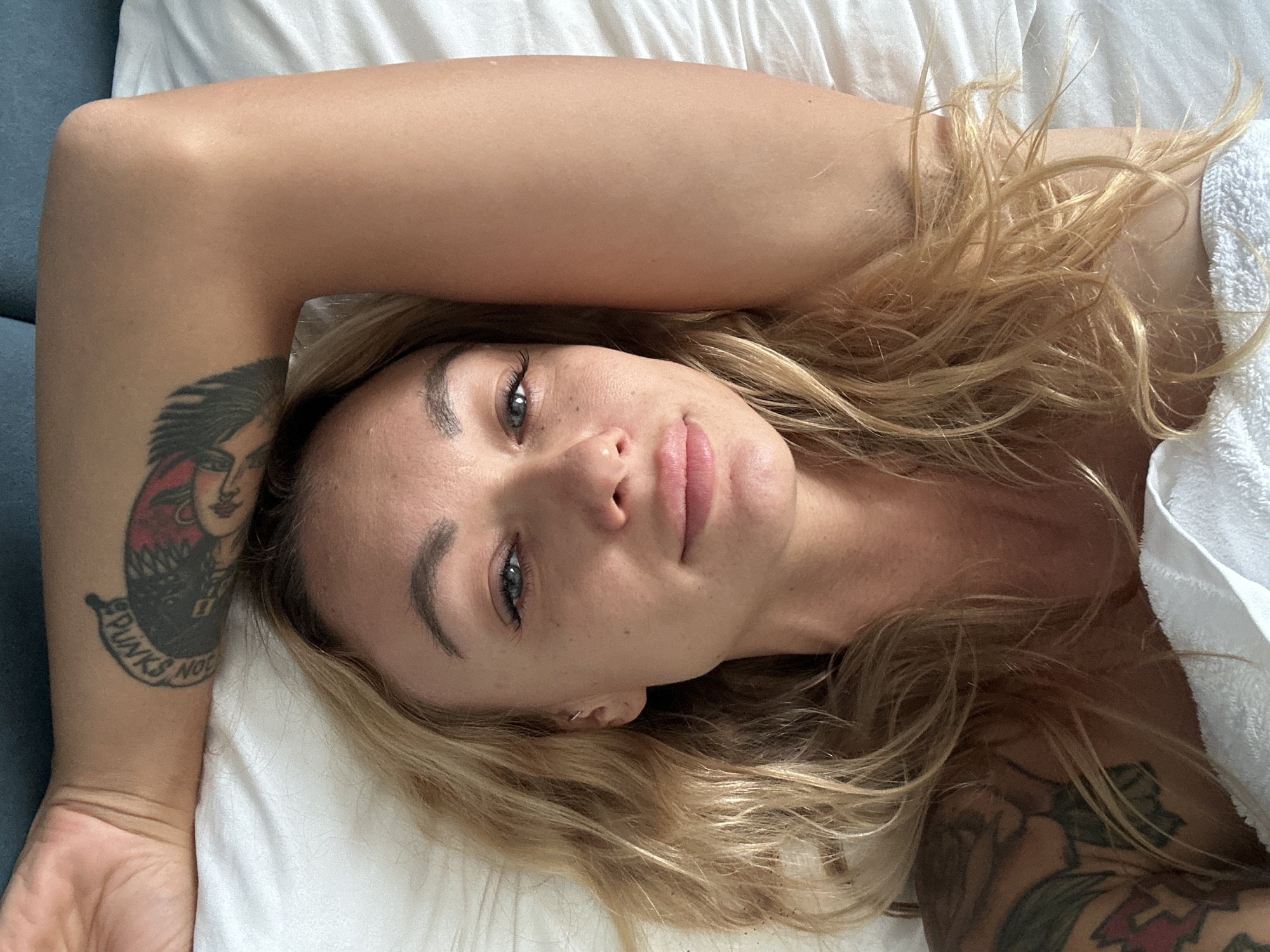 A woman is reclining on a white pillow, her head resting near the top of the frame. She has fair skin, blonde hair, and light-colored eyes. Her left arm is raised above her head, revealing a tattoo of a person on her upper arm with the phrase "PUNKS NOT". Her right arm, also tattooed, is partially visible in the lower right corner of the frame, obscured by a white towel. The woman is facing the camera. A blue surface is seen in the upper left corner of the frame. There is no information on the location of Đồng Nai, Vietnam, in the image.