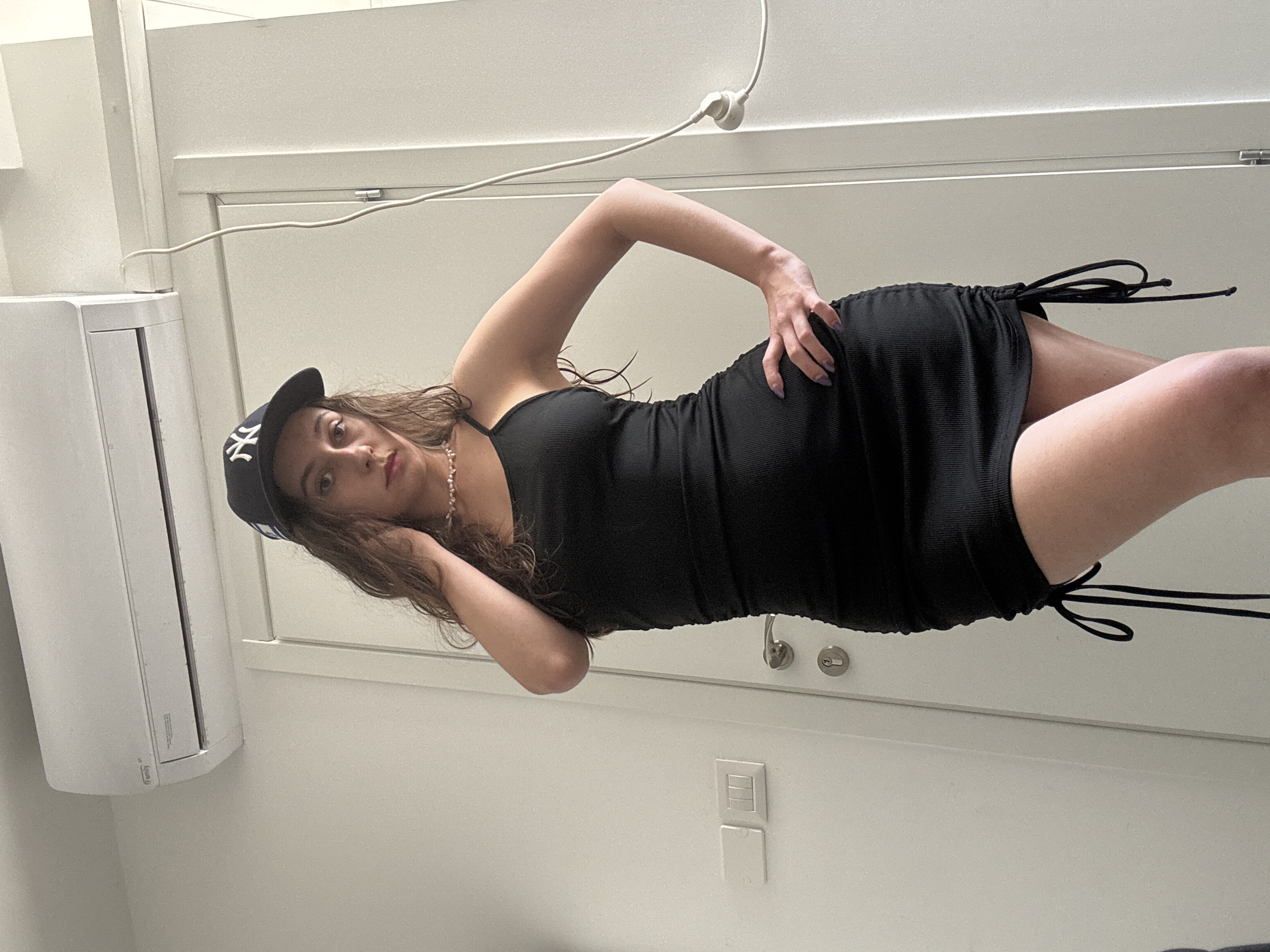 A young woman is depicted lying on her side in an indoor setting, looking directly at the camera. She has long, wavy brown hair and is wearing a black ribbed, sleeveless dress with adjustable side ruching, featuring black drawstrings. A dark baseball cap with a prominent white "NY" logo (New York Yankees) is on her head, and she wears a delicate necklace. One arm is bent with her hand resting on her hip, while her other arm is bent, supporting her head. Her expression is calm and direct, with a hint of red on her lips.
The background consists of plain white walls, suggesting a modern and minimalist room. To her left, an air conditioning unit is mounted, with the word "Family" visible on its side, along with some smaller, less discernible text. A white electrical cable with a plug attached runs across the upper part of the frame, originating from the ceiling or a high wall. On the wall behind her lower body, a light switch panel or outlet is visible. The overall scene conveys a clean, brightly lit environment. Based on the provided context, the location is Ramat Gan, Israel. The image is rotated 90 degrees counter-clockwise.
