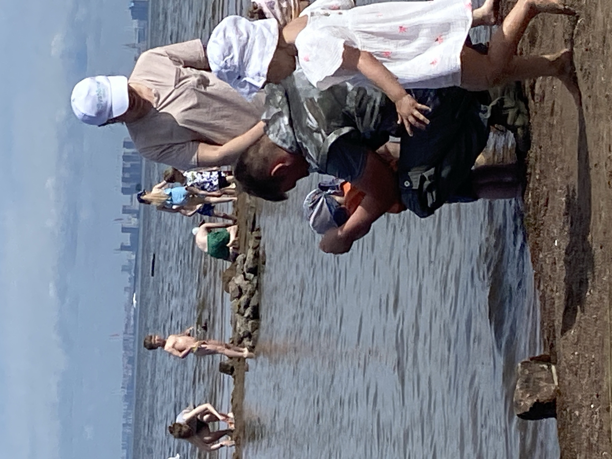 A group of people are on a sandy and rocky shoreline, with the Gulf of Finland in the background. A hazy cityscape is visible on the horizon, likely St. Petersburg. In the foreground, a man wearing a camouflage vest and dark pants is kneeling, holding a small child in a blue cap. Beside them, a young girl in a white dress and hat stands. Further back, several other people, including children, are scattered along the water's edge, some interacting with the water or building with sand and rocks. The water is calm, with gentle ripples. The sky is overcast with some blue patches visible.