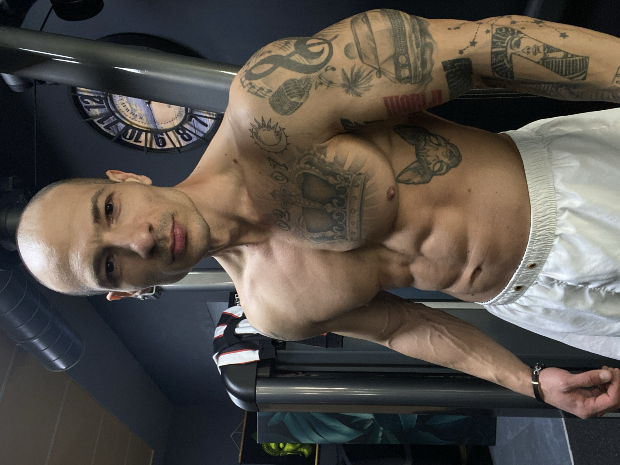 A muscular, bald man with tattoos is standing indoors. He is wearing white shorts and a black bracelet on his left wrist. He is in front of a piece of gym equipment with a black and gray bar. Behind him is a gray wall with a decorative clock on it. A treadmill with artwork on the lower panel is partially visible behind him. The exact location in Pleven, Bulgaria, is indeterminable from the visual information provided.