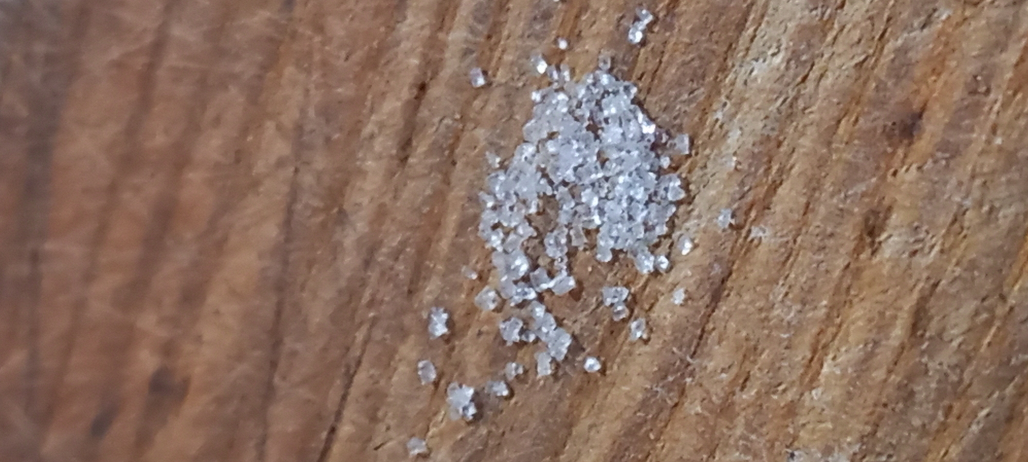 The image displays a close-up, high-angle view of a small cluster of white, crystalline granules resting on a wooden surface. The granules, likely coarse salt or sugar, are translucent and irregularly shaped, with facets that catch and reflect light, creating a sparkling appearance. They are concentrated in the upper right portion of the frame, with a few individual crystals scattered nearby.
The background consists of a warm, light to medium brown wooden surface, possibly a kitchen counter, cutting board, or tabletop. The wood features distinct grain patterns running diagonally, showcasing its natural texture and imperfections. The surface appears slightly worn, contributing to its rustic character.
No specific activity is shown, but the presence of the granules suggests a moment related to food preparation or a minor spill. The lighting illuminates the details of both the textured wood and the reflective crystals. There are no people, visible text, or direct environmental cues within the image that indicate a specific location. The image's focus is on the granular texture of the crystals against the natural wood background. While the context of Kryvyi Rih, Ukraine, is provided, it is not visually verifiable from the image content.