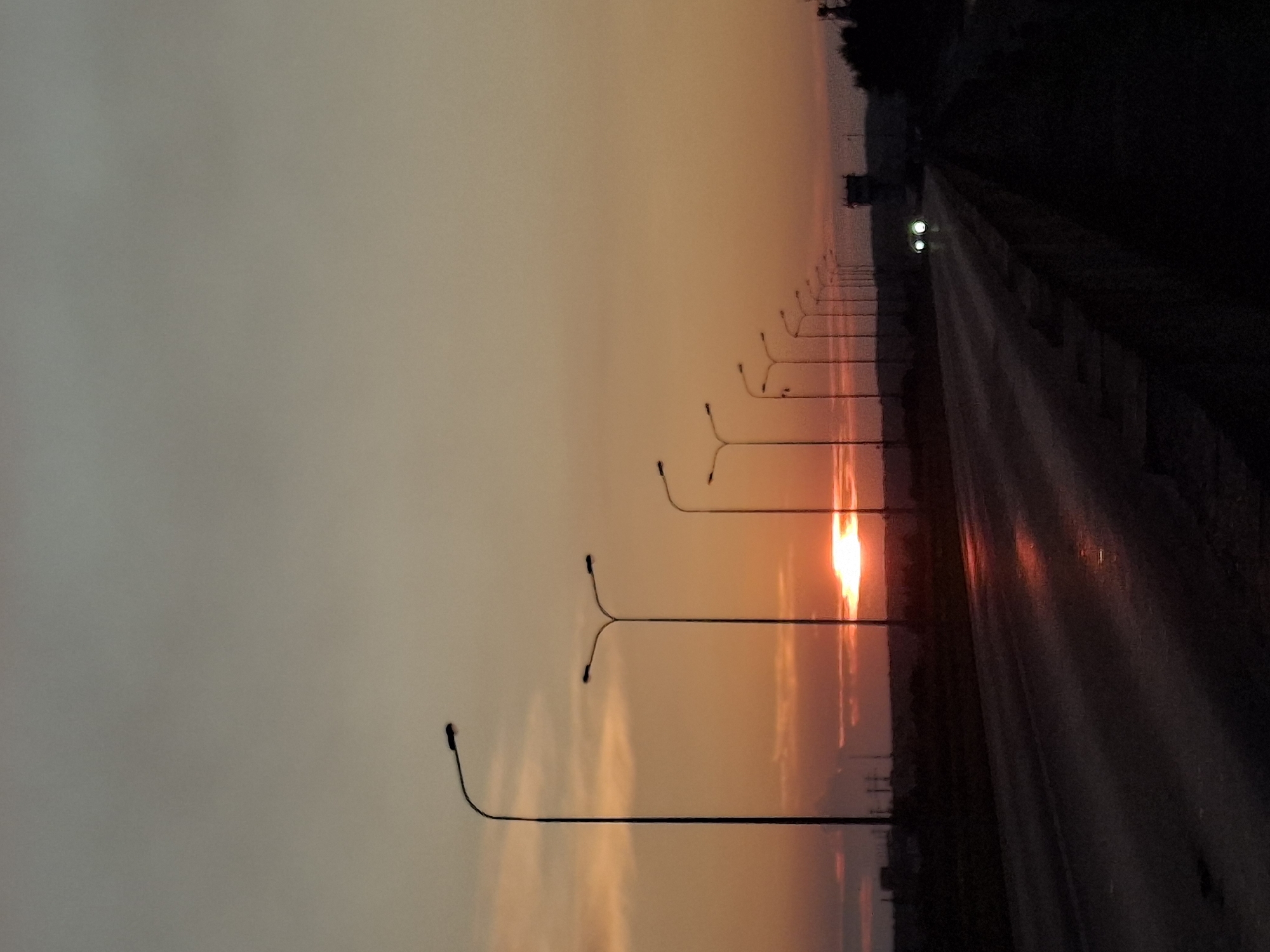 A highway at sunset in Chaikovsky, Russia. The sky is a gradient of pale gray to warm orange and red as the sun sets near the horizon. A line of streetlights with dark poles and fixtures lines the right side of the highway. The highway itself is dark and wet, reflecting the orange glow of the sunset. In the distance, two car headlights are visible on the highway. The surrounding landscape is dark and indistinct, with silhouetted trees and structures.