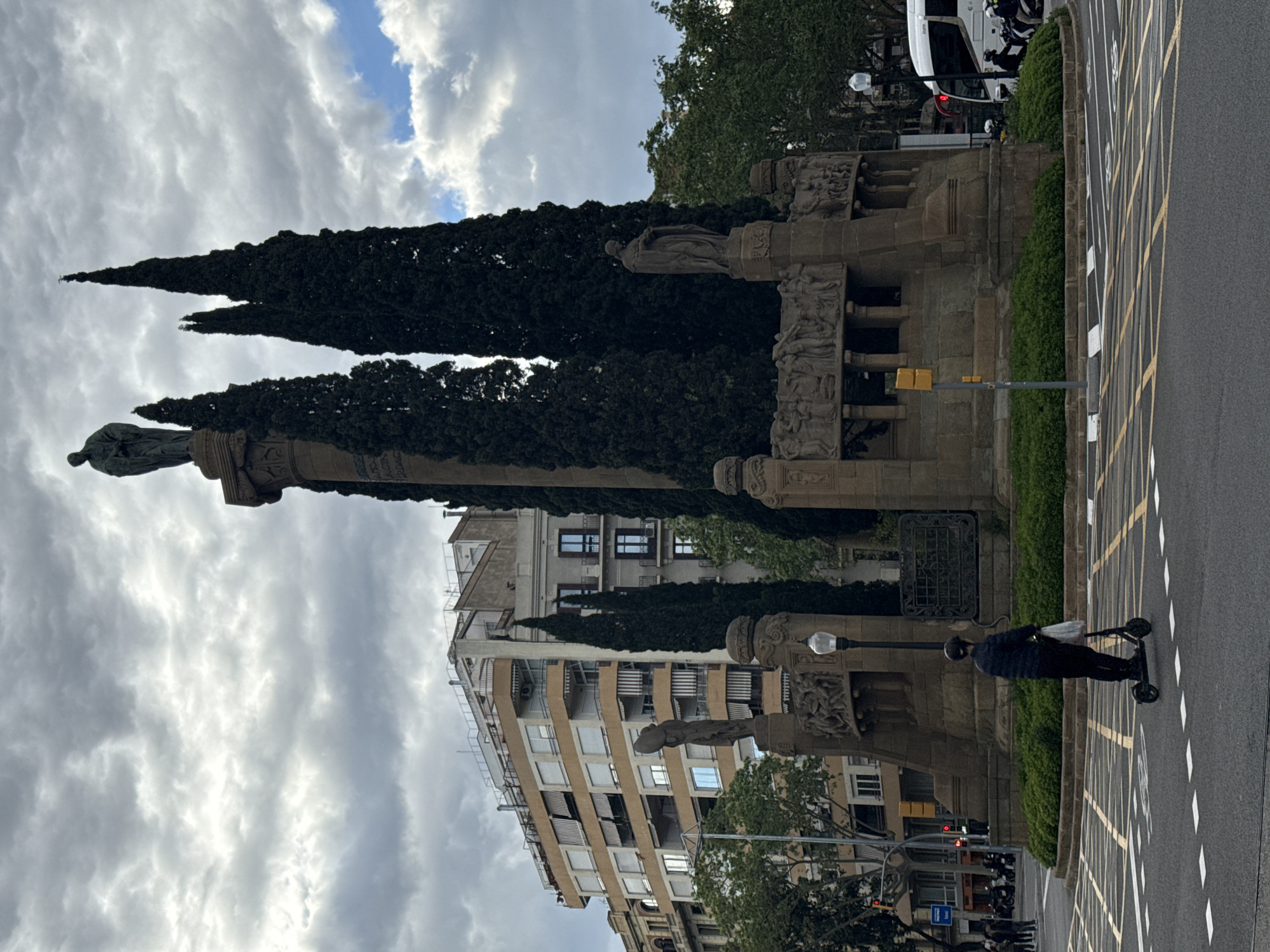 The image depicts a street scene in Barcelona, Spain, with the Monument a Colom (Columbus Monument) prominently featured. The monument, topped with a statue of Christopher Columbus, stands tall amidst slender cypress trees. Intricate carvings adorn the monument's base.
In the foreground, a person rides an electric scooter along a city street marked with yellow and white cross-hatchings. A lamppost stands near the scooter rider, blending in with the monument's stonework.
Background buildings with windows and balconies add to the urban landscape. The sky is overcast, with layered clouds. A white vehicle with flashing lights is visible parked at the road edge adjacent to the monument. Pedestrians can be seen in the distance at a traffic signal.