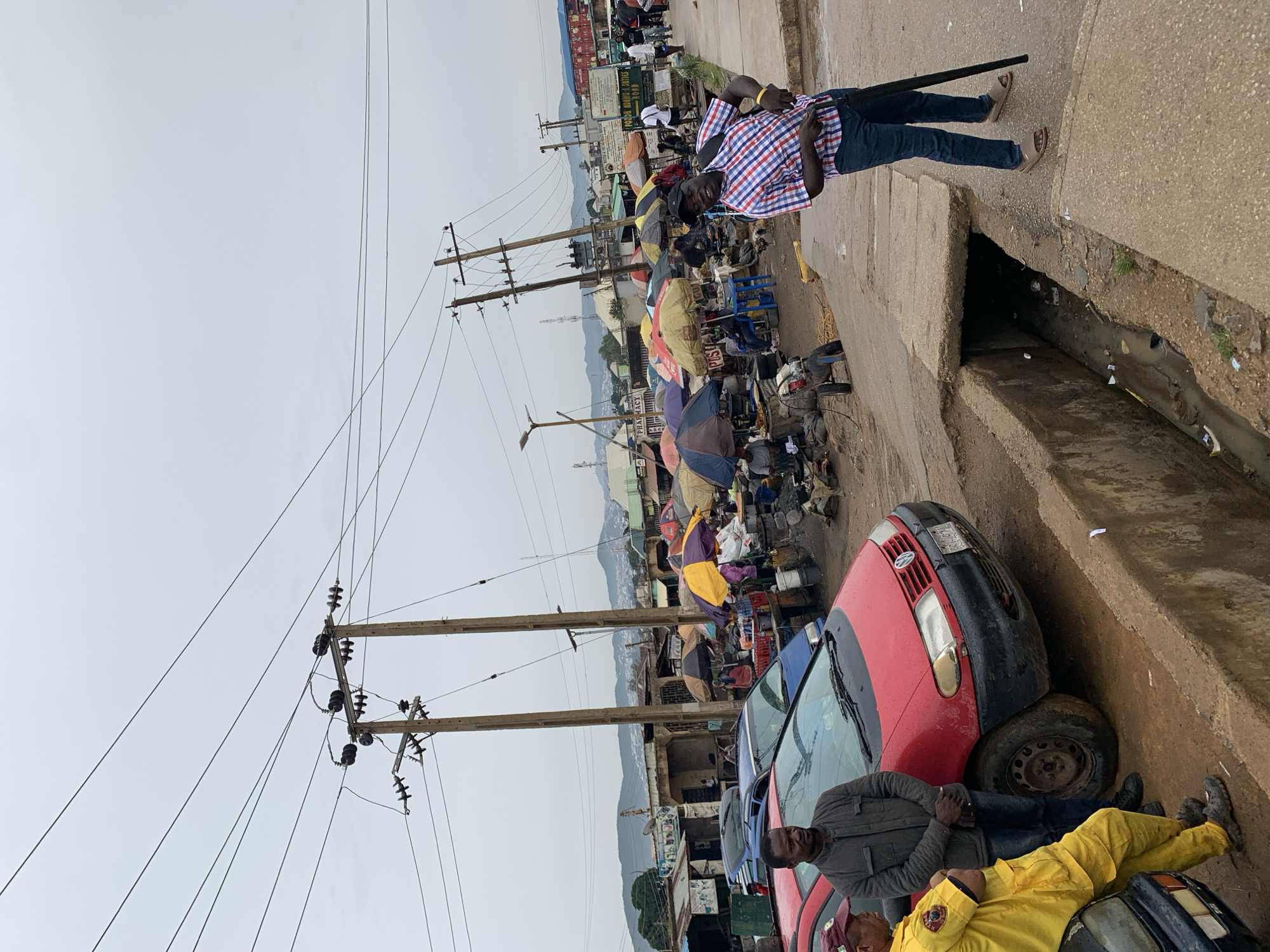 The image depicts a street scene in Rafin Par, Nigeria. Two men are near a row of parked cars, one red, and one blue. One of the men is wearing a yellow outfit and is lying on the front of a car. The other man is wearing a grey jacket and is standing next to the man lying on the car. In the background, there are many people, market stalls with umbrellas, and power lines with poles. A man in a red and white checked shirt and jeans is standing on a concrete walkway. A trench runs along the side of the road. In the distance, there are buildings and mountains. The sky is overcast.