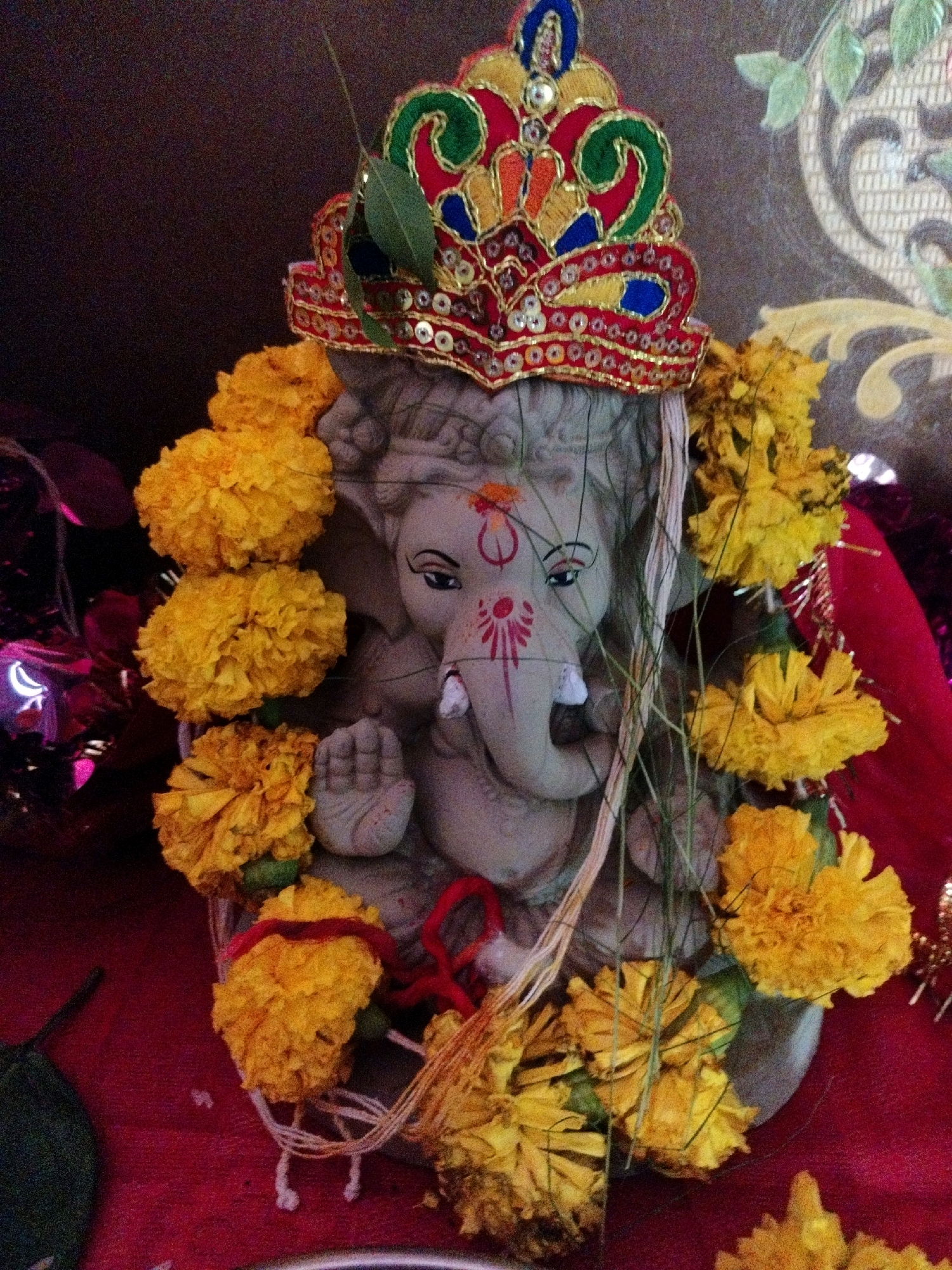 The image shows a statue of Ganesh, a Hindu deity, adorned with yellow marigolds and a decorated crown. The statue is placed on a red cloth. The background is a dark, textured wall with some decorative elements. There are no people or actions depicted in the image. The location is unknown and cannot be determined from the image.