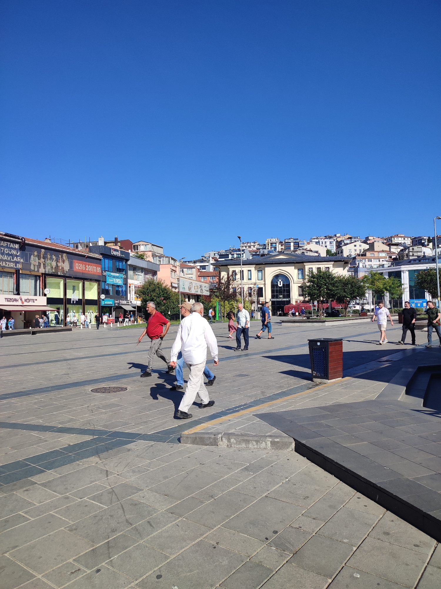 The image shows a sunny day in a public square in Üsküdar, Turkey. The sky is clear and bright blue. In the foreground, several people are walking across the paved square. One man in a white shirt and light-colored pants is walking towards the viewer. To his left, a man in a red shirt and grey pants is walking away from the viewer. Other individuals are scattered across the square, some walking alone, others in small groups.
The square is lined with buildings. On the left, there are commercial establishments with storefronts displaying advertisements and mannequins. Further back, more buildings rise up the hillside, characteristic of urban architecture in the region. In the center of the background, a large, light-colored building with a prominent arched entrance stands out. Trees are planted in the square and around the buildings. A dark-colored trash receptacle is visible in the middle right of the image. The paving stones of the square are laid in a geometric pattern, with some darker bands indicating pathways or design elements. Shadows cast by people and buildings suggest the sun is high in the sky.