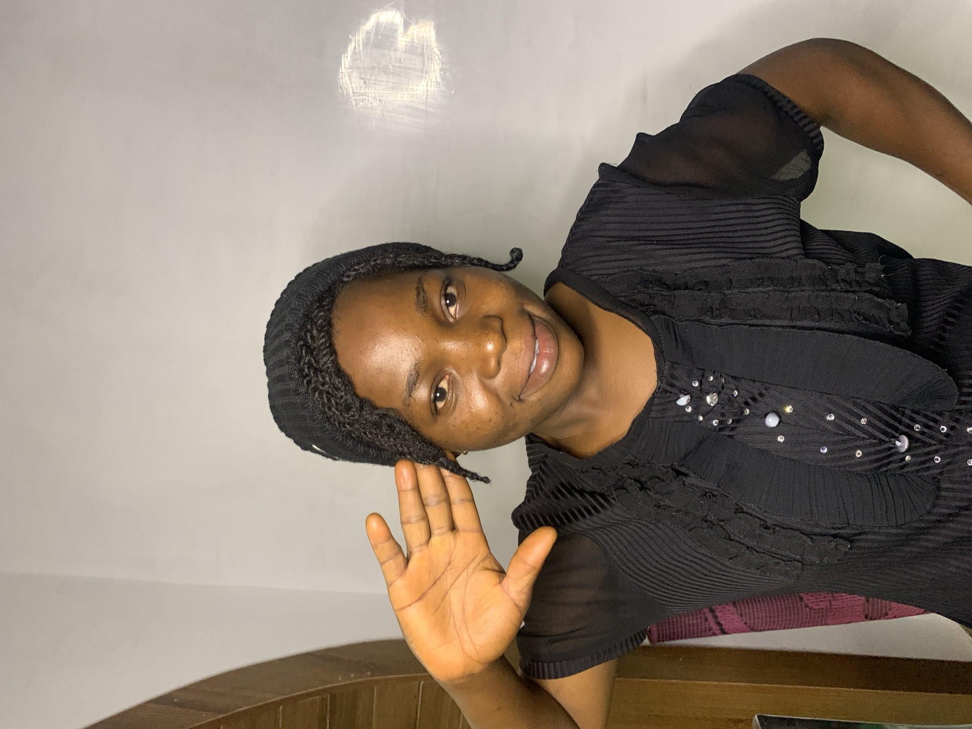 A medium shot shows a Black woman with dark skin and braided hair, wearing a black headwrap and a black top with ruffled detailing and embellishments on the neckline. She is smiling and holding her right hand up, palm facing the camera, near her forehead. The background is a plain white wall with a subtle reflection. A wooden door frame is visible on the left side of the image. This image was taken in Jos, Nigeria.