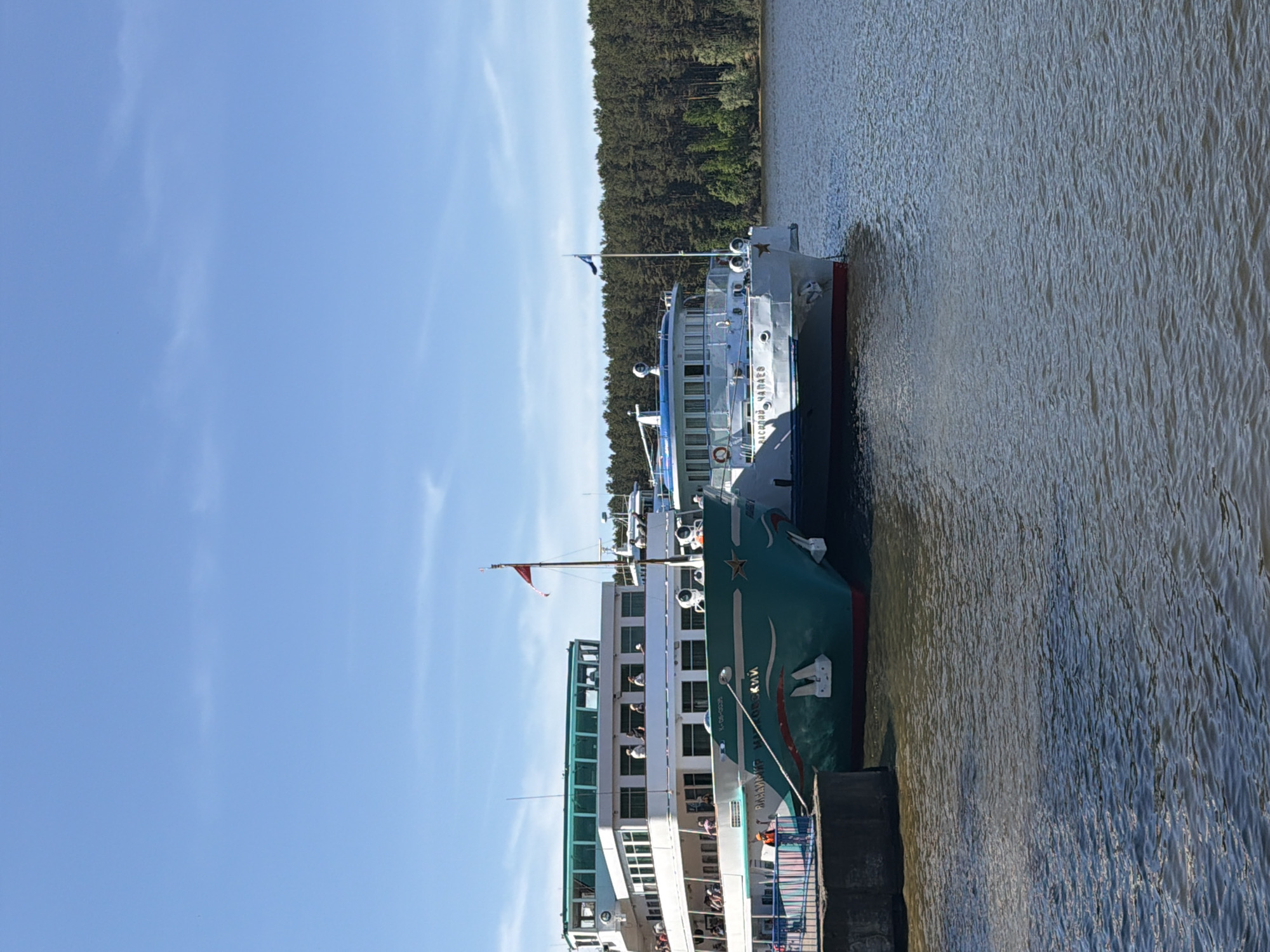 The image depicts two passenger ships docked at a pier. The ship closer to the viewer has a green hull, white superstructure, and multiple decks filled with people. A red flag flies from a mast. The second ship has a white and gray hull. The background features a dense forest and a blue sky with light clouds. The water is choppy. The image likely shows the city of Chaikovsky, Russia, due to the architecture of the boats being of Russian descent.