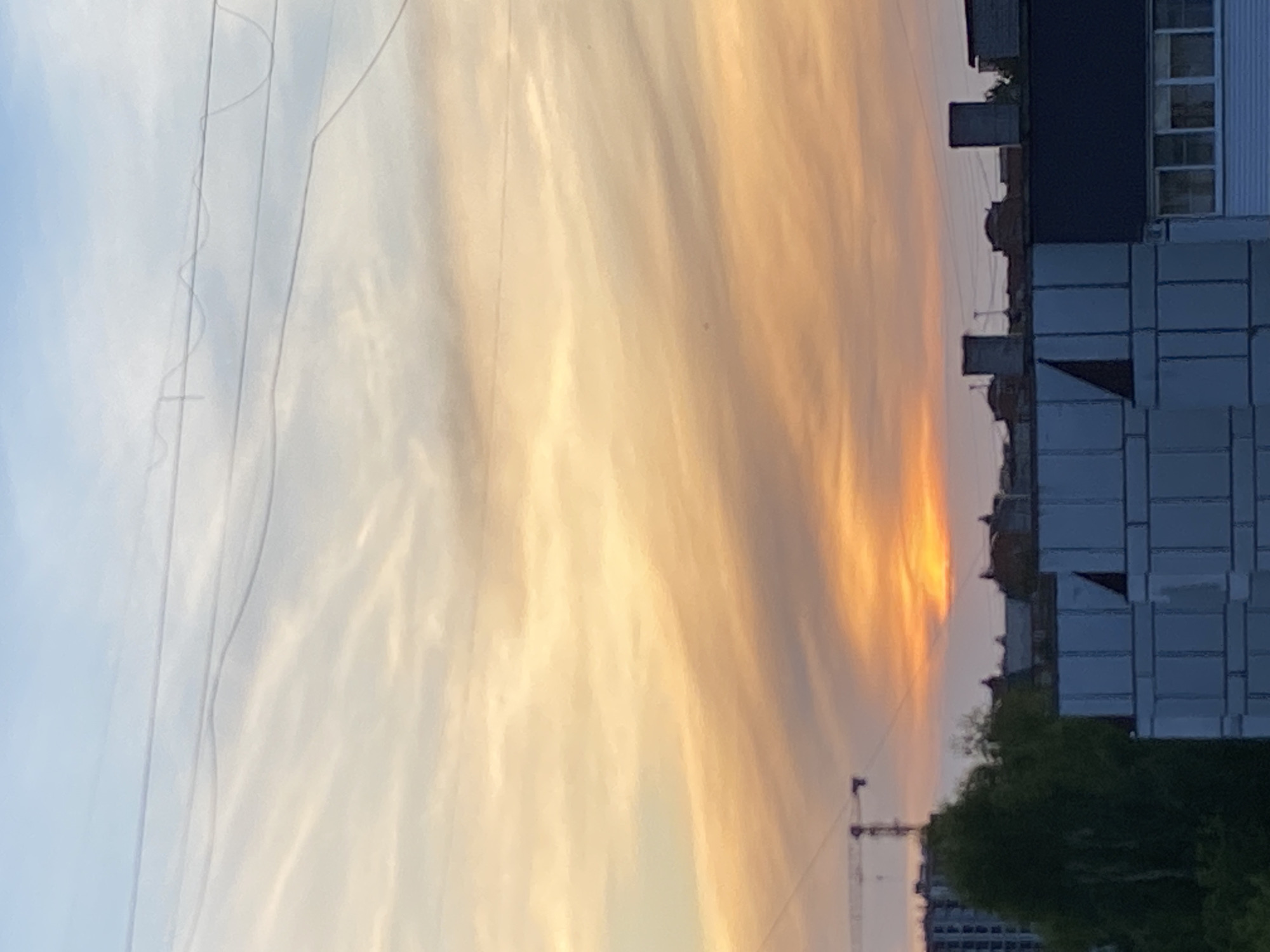 Here is a detailed description of the image:
The image captures a sunset in what appears to be a residential or urban area. The sky is filled with clouds exhibiting warm, golden and orange hues, indicative of a sunset or sunrise. Utility lines are visible against the backdrop of the sky, running horizontally across the frame.
On the right side of the image, there is a tall building with multiple windows and architectural features. The building's color is a mix of light and dark shades, possibly due to the angle of the light and the shadows it casts.
Below the building, there are trees with green foliage, which provide a contrast to the building and the sky. In the distance, there's a construction crane, hinting at ongoing construction or development activities in the area. The horizon line shows other buildings of varying heights, suggesting this is an urban setting. Based on the prompt, this image is likely in the city of Chaikovsky, Russia.