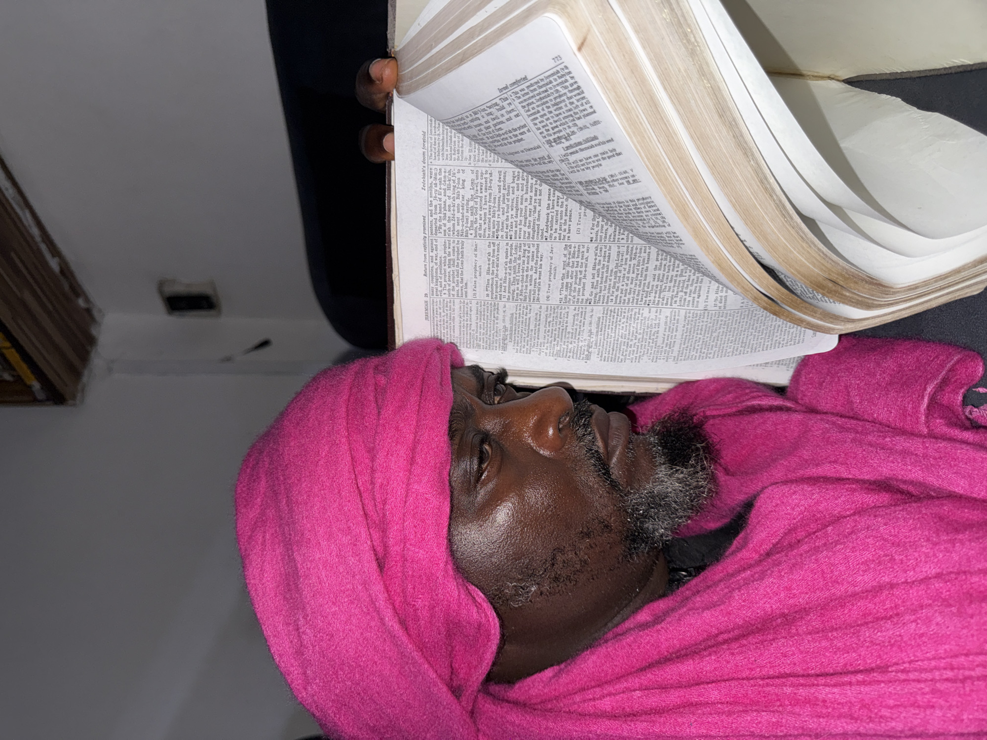 A middle-aged man with dark skin, graying beard, and eyes open, is lying down covered in a pink cloth. He is holding a thick, open book with thin, white pages directly over his face. The book is turned to Jeremiah 29. The environment is inside an underexposed room, with a wall, part of a piece of furniture, and an electrical outlet visible. The image's location in the city of Ado, Nigeria, is not verifiable.