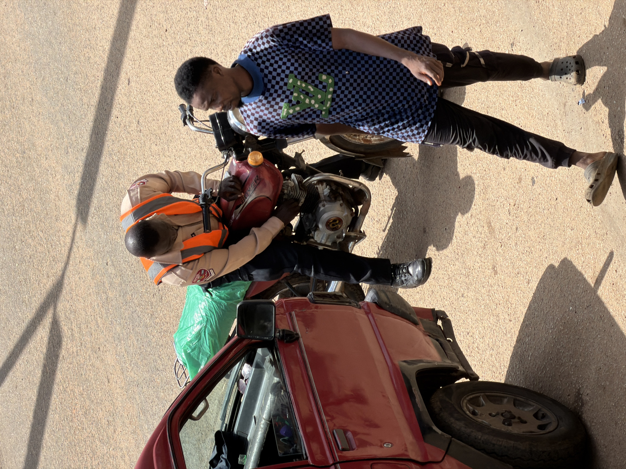 Two young men are interacting with a motorcycle and a red vehicle in an outdoor setting. One man, wearing a checkered blue and black shirt and black pants, is seated on the motorcycle, leaning forward. He has dark skin and short, dark hair. The second man, dressed in a tan uniform with an orange safety vest, is also positioned on or near the motorcycle, appearing to be working on it. He is also wearing dark pants and black shoes. A bright green plastic bag is visible near the motorcycle. The red vehicle, a pickup truck, is partially visible with its door open and a visible side mirror. The ground appears to be a paved surface, possibly asphalt, with visible shadows indicating it is daytime, likely late morning or early afternoon given the length and angle of the shadows. The scene appears to be on a street or roadside in Bukuru, Nigeria. The overall impression is one of a mechanical issue or repair being addressed on the motorcycle. There is no visible text in the image.