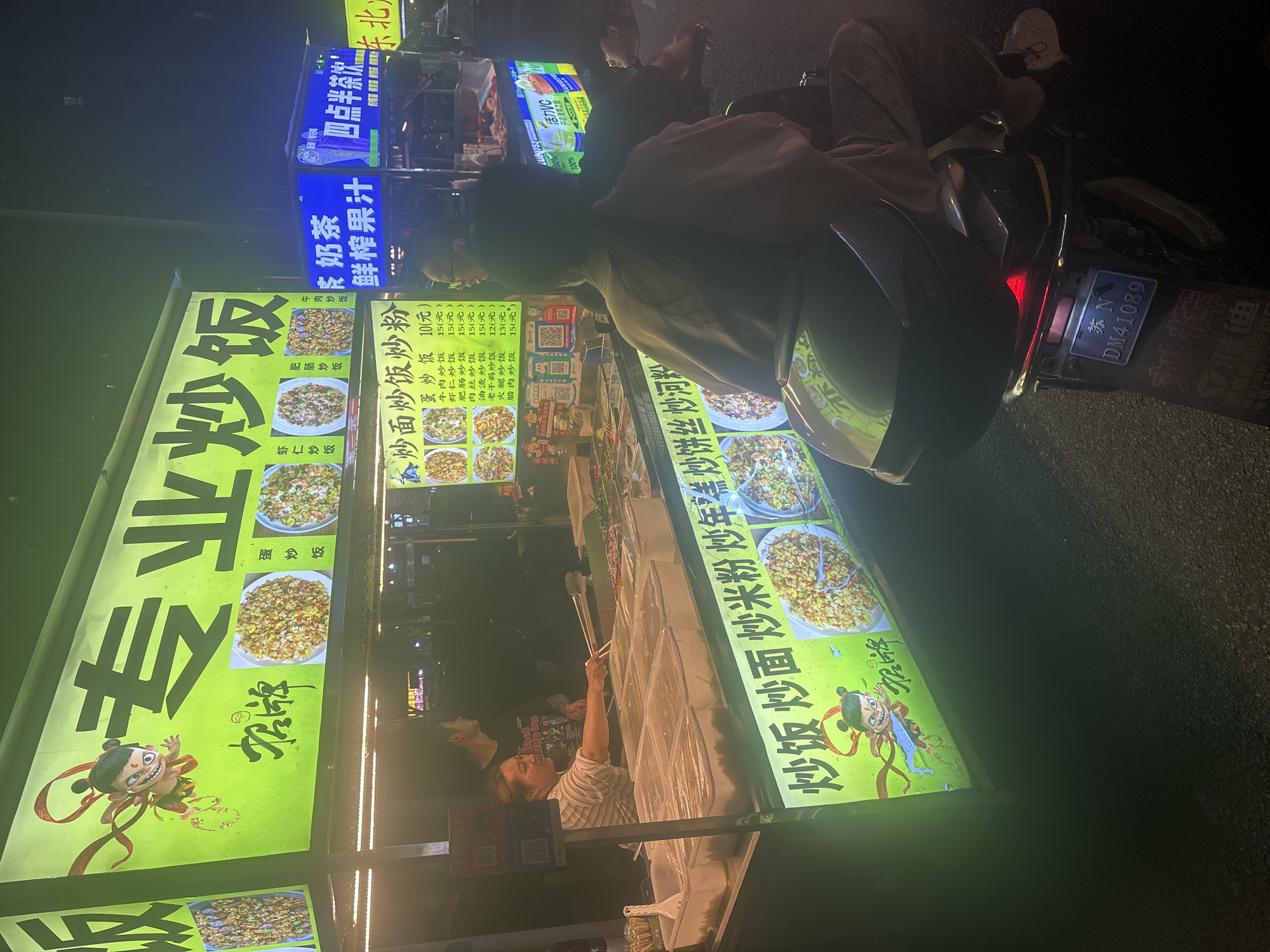 In Suzhou Industrial Park, China, a nighttime scene unfolds at a bustling street food stall specializing in fried rice. A brightly lit green sign, adorned with a cartoon character and Chinese calligraphy, announces "Specialty Fried Rice." Below, a display showcases various fried rice dishes, their vibrant colors tempting passersby. A female vendor, her face illuminated by the neon glow, expertly works behind the counter, her tongs poised over an array of ingredients. To her left, a man, presumably a customer, stands, observing the preparation. In the foreground, a person sits on a scooter, facing away from the camera, their presence adding to the dynamic atmosphere. The overall impression is one of lively urban commerce, where delicious food and human interaction converge under the shroud of night.
