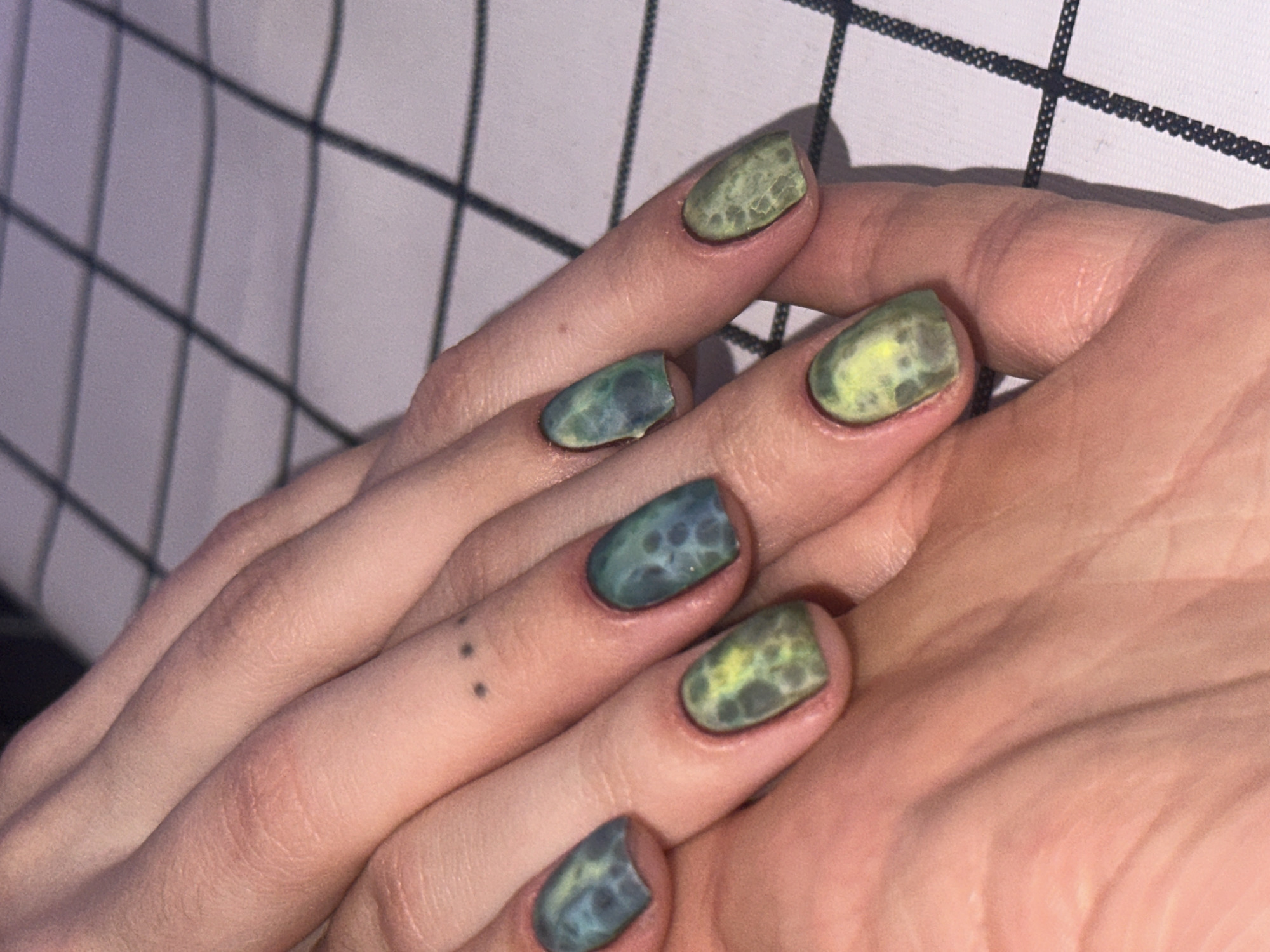 The image shows a pair of human hands with painted nails. The nails have a mottled pattern in shades of green and blue. There is a pattern visible in the background that appears to be a white surface with a black grid. There is a tattoo of three small dots on one of the fingers.
