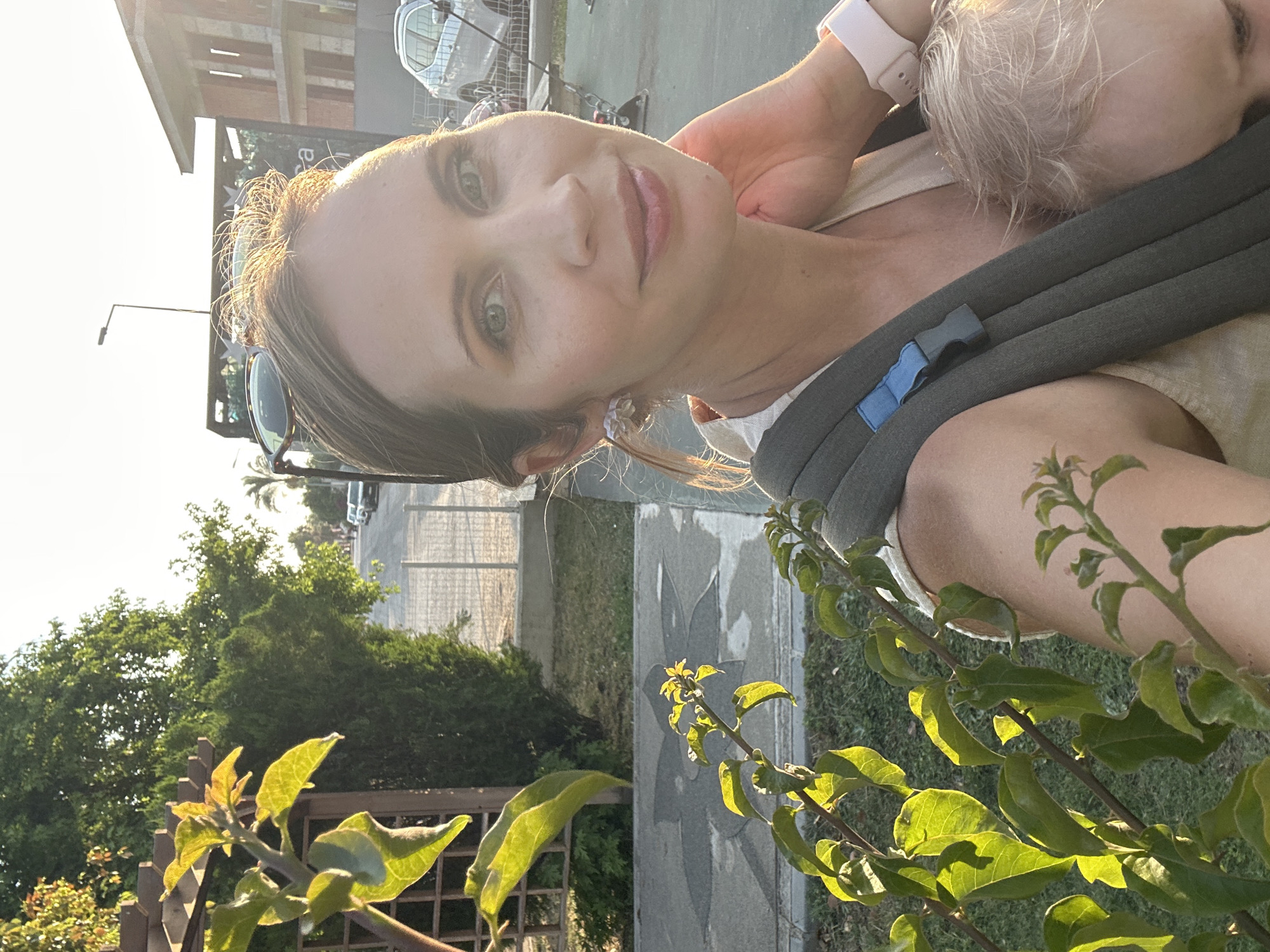A selfie is taken from a woman carrying a baby in a sling, in Fethiye, Turkey. The woman is looking towards the camera, with her hand on her cheek. The baby, who is wearing a light-colored shirt, is visible in the sling. They are outdoors, with a street and buildings in the background. There is a plant in the foreground. The sun is setting, casting a warm glow on the scene. The woman is wearing earrings and a watch.