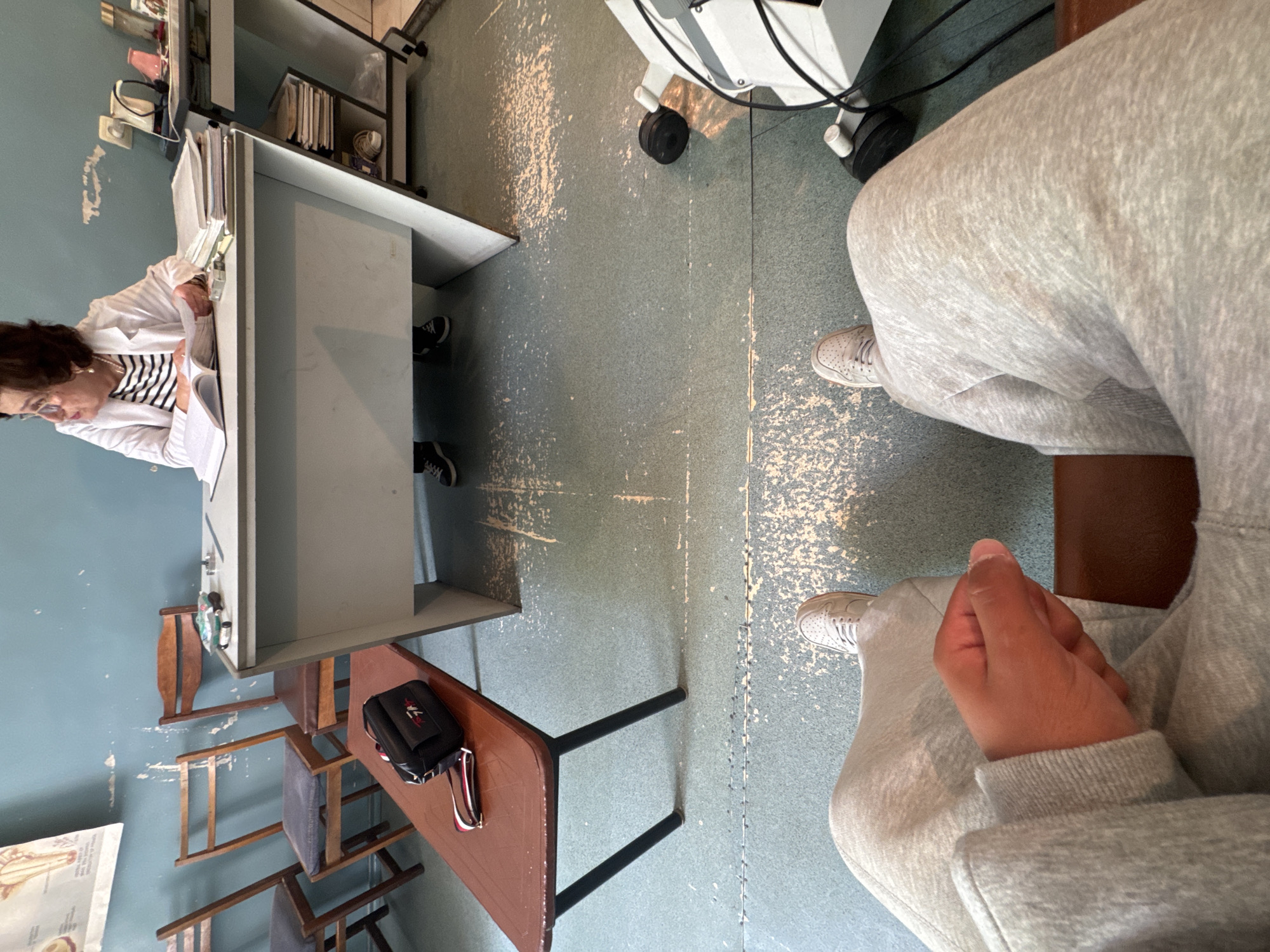 The image depicts the interior of what appears to be a medical office or clinic in Yerevan, Armenia. A female doctor with brown hair and glasses is sitting at a desk, reviewing documents. She is wearing a white coat over a striped shirt. The desk contains documents and medical supplies. The room is sparsely furnished with a metal desk and chairs. There is medical equipment on wheels. The floor has a pale green finish with noticeable wear and tear. A person wearing gray sweatpants and tennis shoes sits in the foreground with their hands clasped. A purse sits on the edge of the table, along with keys and glasses. The room has pale blue walls. An anatomical chart is on the wall.