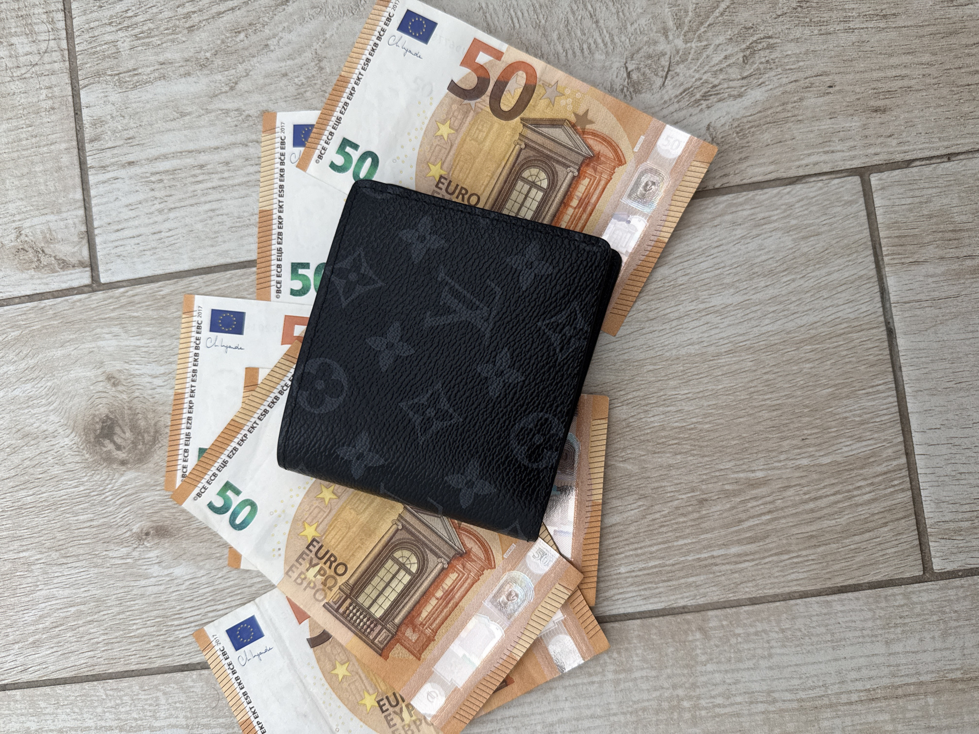 A black wallet with a Louis Vuitton monogram pattern lies on a pile of 50 Euro banknotes. The banknotes are spread out on a light brown, wood-patterned tiled floor. The wallet is positioned in the center of the image, partially covering the banknotes. Some of the banknotes are folded. The scene is lit from above, casting subtle shadows. The image appears to be a top-down shot. There is no indication of the city of Burgas, Bulgaria.