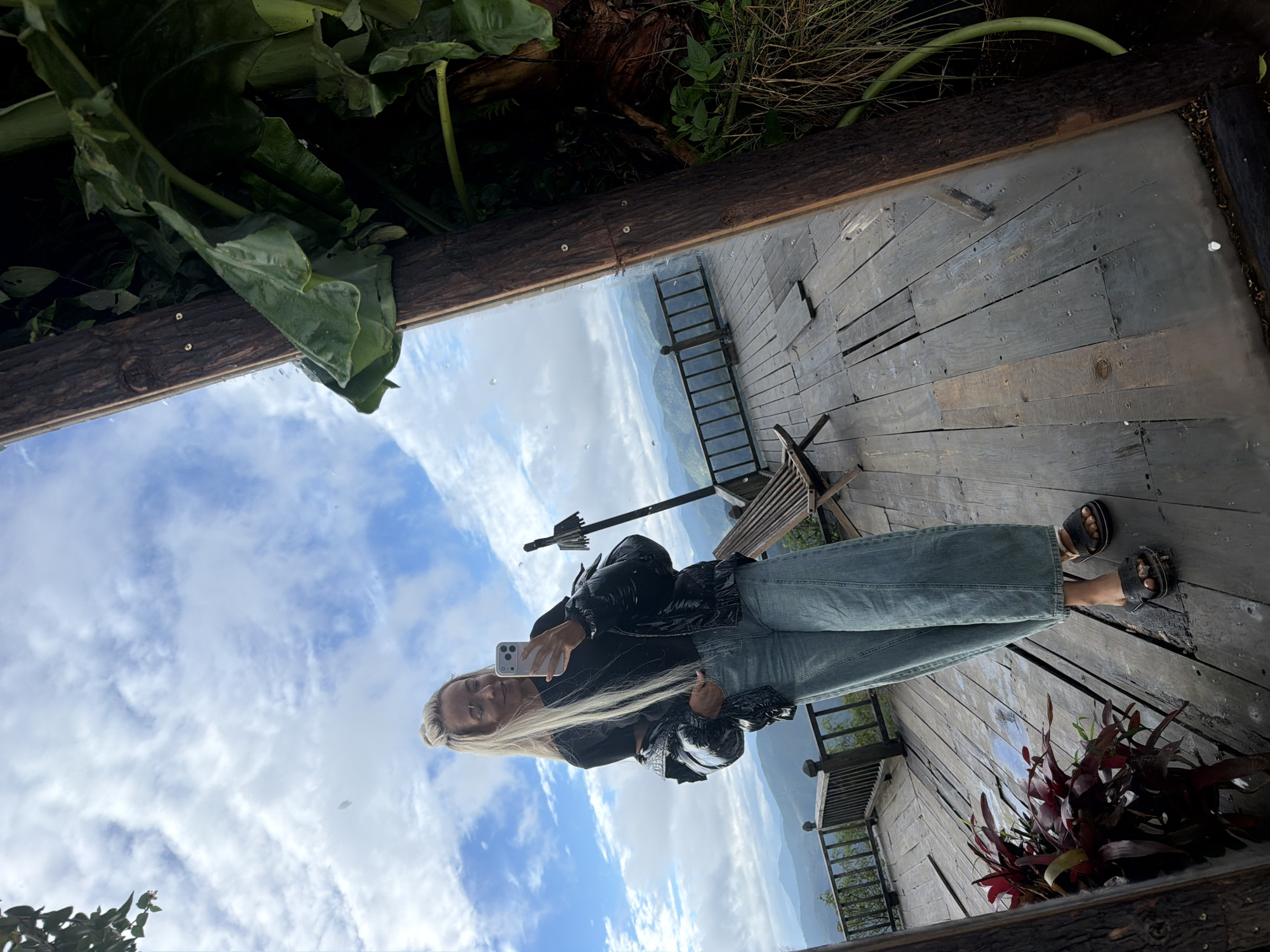 A full-body selfie is taken by a young woman with long blonde hair pulled up in a bun and bangs, wearing glasses, a black t-shirt, a shiny black puffer jacket, light blue wide-leg jeans, and black sandals. She is standing on a wooden deck with a railing overlooking a mountainous landscape with a cloudy sky. A large mirror reflects the scene. To the right of the mirror, there is lush green foliage and a wooden post. In the foreground on the left, there is a plant with dark red leaves. A wooden chair is visible on the deck behind the woman. The location is identified as Phát Chi, Vietnam.