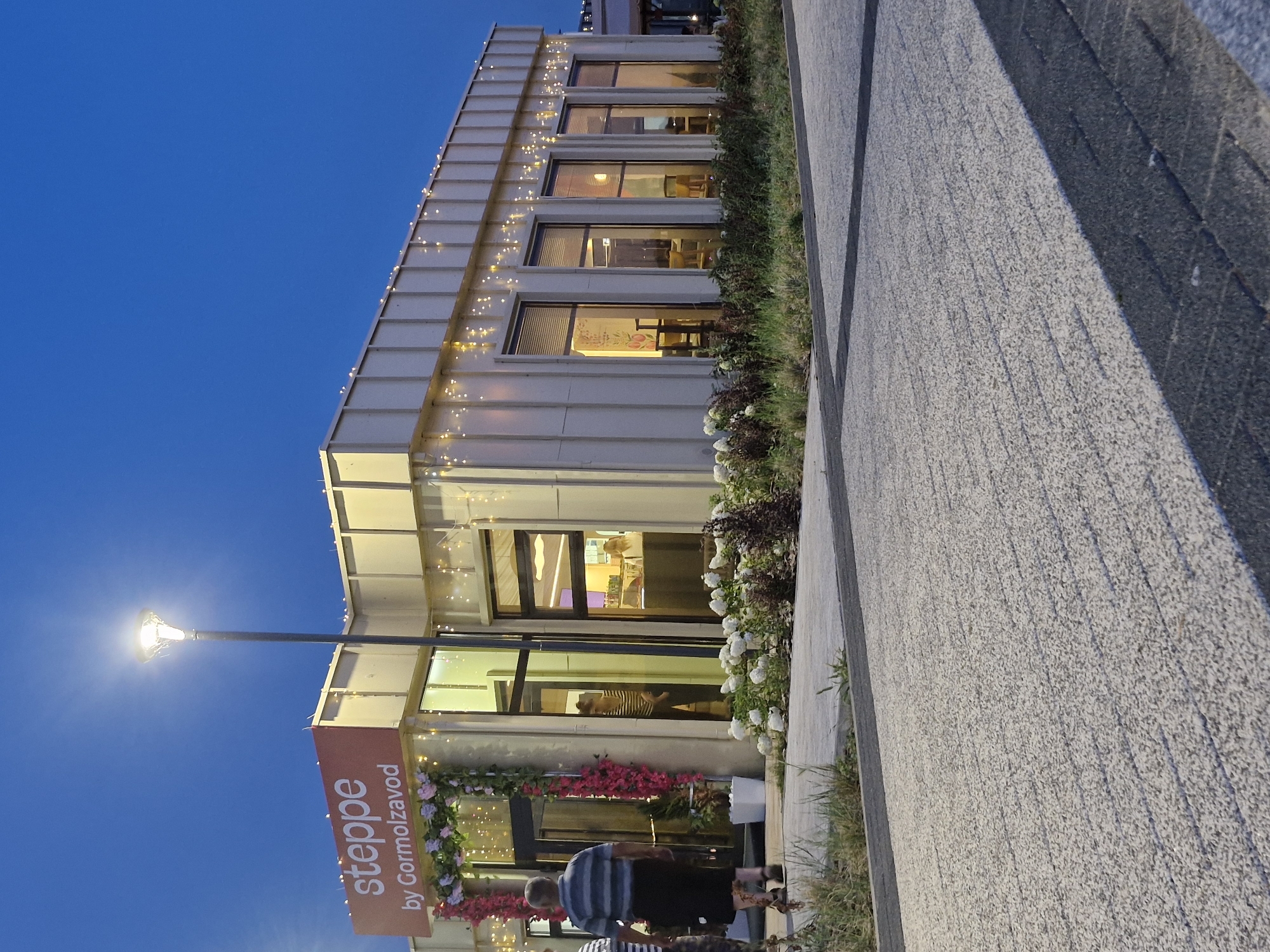 A vertical, eye-level medium shot captures the exterior of a modern building at dusk, with a clear, deep blue sky above. The building features a light-colored, modular facade adorned with a string of small, warm-toned lights running along its upper edge. Large windows reflect the interior lights and the twilight sky, offering glimpses of sparsely furnished rooms with yellow chairs and tables.
In front of the building, a pathway paved with textured gray concrete runs diagonally across the frame. To the left of the pathway, a vibrant orange banner with white text reads "steppe by Gormolzavod." The entrance to the building is decorated with a floral archway featuring pink and purple flowers.
A single streetlamp on a dark pole illuminates the scene from the left, casting a bright glow. The ground around the building is lined with grass and some darker foliage. Two people, partially visible, are walking away from the camera on the pathway. One person wears a blue and white striped shirt and dark pants, while another, in the foreground, wears a similar striped shirt and dark shorts. The overall atmosphere is calm and inviting, suggesting the early evening of a pleasant day in Kokshetau, Kazakhstan.