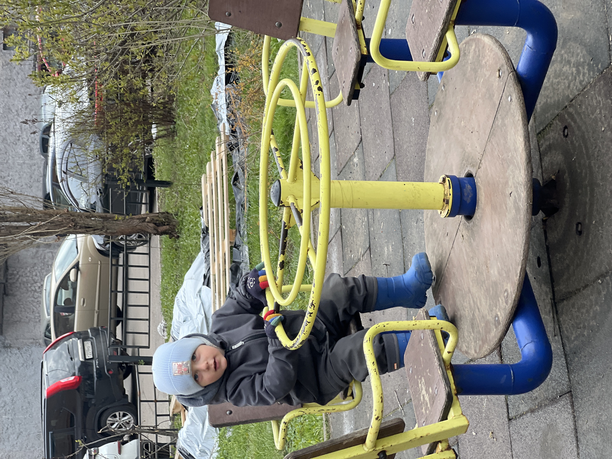 The image captures a young child, appearing to be a toddler or preschooler, sitting on a playground merry-go-round. The child wears a dark grey hooded jacket, a light blue knitted hat, and bright blue rubber boots with colorful striped gloves, indicating cool weather. Their expression is neutral as they look directly at the viewer, holding onto the worn yellow handlebars of the merry-go-round. The equipment features wooden seats and a central yellow pole, with much of its yellow paint chipped, exposing the metal beneath. The surrounding ground is paved with grey tiles.
In the background, signs of early spring are visible with trees and bushes bearing sparse, fresh green leaves. Beyond the foliage, an urban or suburban street is visible with several parked cars. A prominent black SUV-type vehicle displays a partially legible Russian license plate, "P000TP790". A beige sedan is parked further back, and hints of another white vehicle are visible. Alongside the street or path, there are wooden planks and white tarps or sheeting, suggesting ongoing work or stored materials. The scene depicts an outdoor playtime moment during daylight hours in Mytishchi, Russia, set in a public or residential area.
