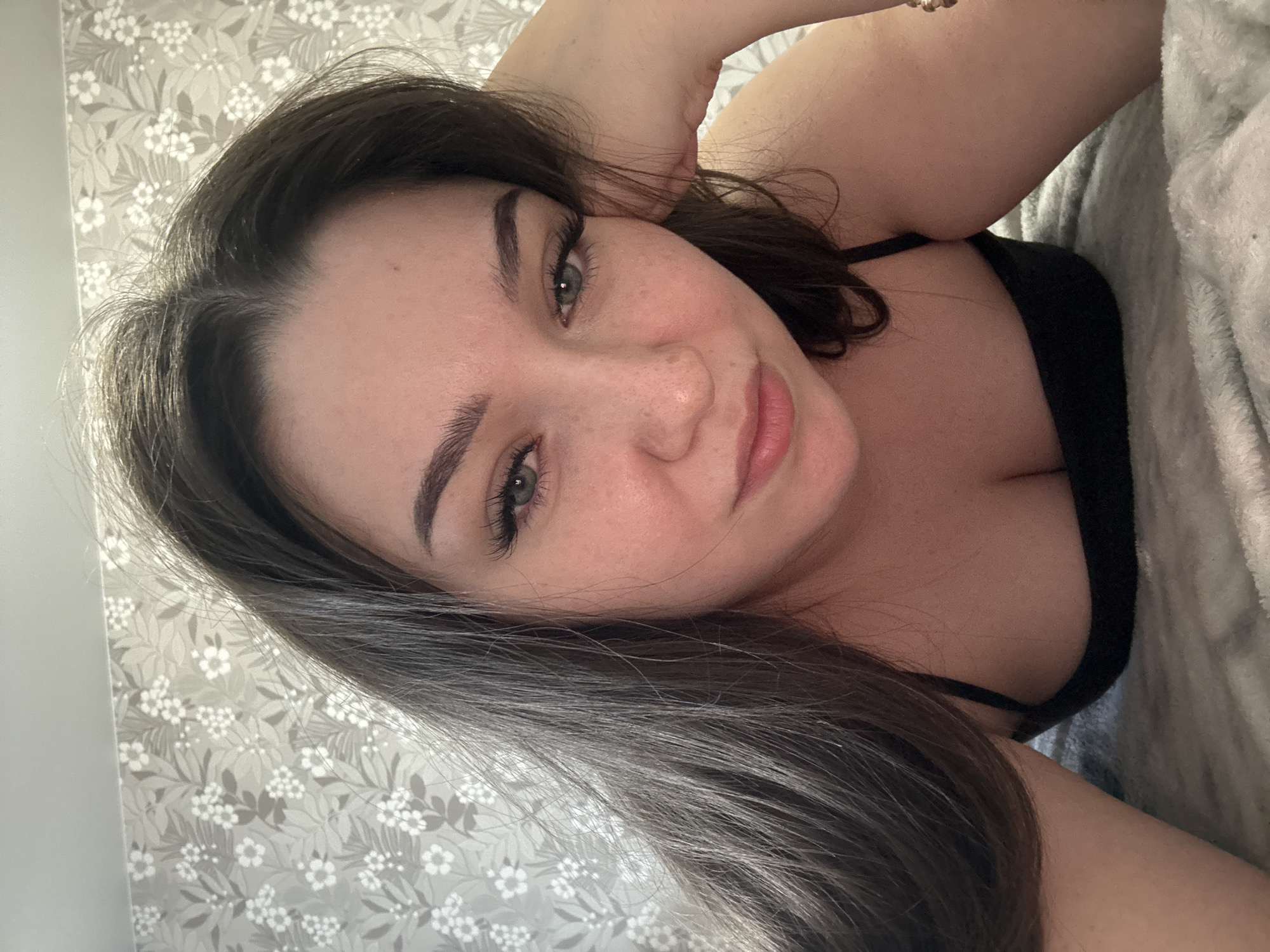 This is a portrait-oriented selfie of a young woman with dark brown hair, lying on her side. She is wearing a black top. Her left arm is bent, with her hand resting behind her head. The background features a floral wallpaper with a light gray and white pattern. To her right, a gray textured blanket is visible. The lighting suggests it might be daytime, possibly indoors. Her expression is relaxed, with a slight smile. There is no visible text in the image.