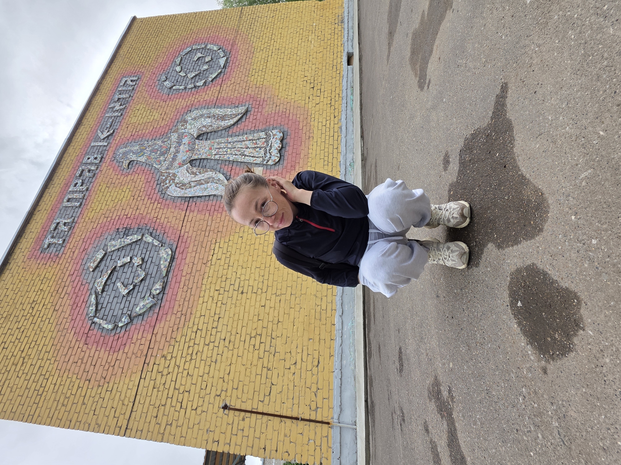 A full shot taken outdoors, shows a fair-skinned woman crouched on a gray asphalt surface, with a yellow brick wall behind her. The wall, positioned on the left, features a mosaic design of an eagle and circular shapes, possibly indicating a crest or logo. In the background, a gray, overcast sky is visible. The woman is wearing circular glasses, a dark-colored jacket, and light-gray sweatpants, paired with light-colored sneakers. There is a puddle of water to the woman's right. The location is the city of Unknown, Unknown.