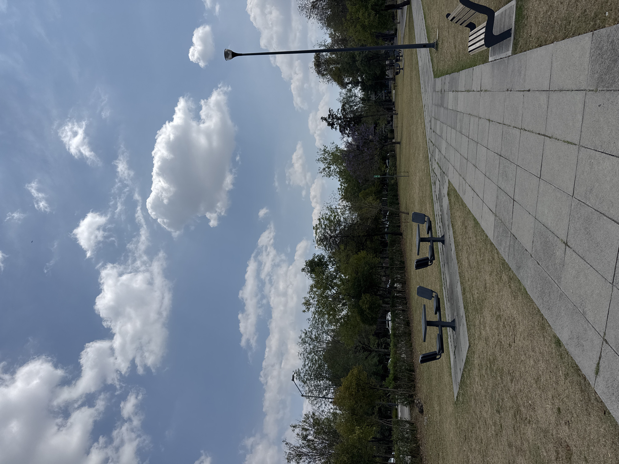 The image shows a park landscape with a tiled walkway. There are benches along the walkway and exercise stations further into the park on a grassy area. The sky is partially cloudy. Based on the architecture and flora, it is likely located in the city of Puebla, Mexico. No people are visible in the image.