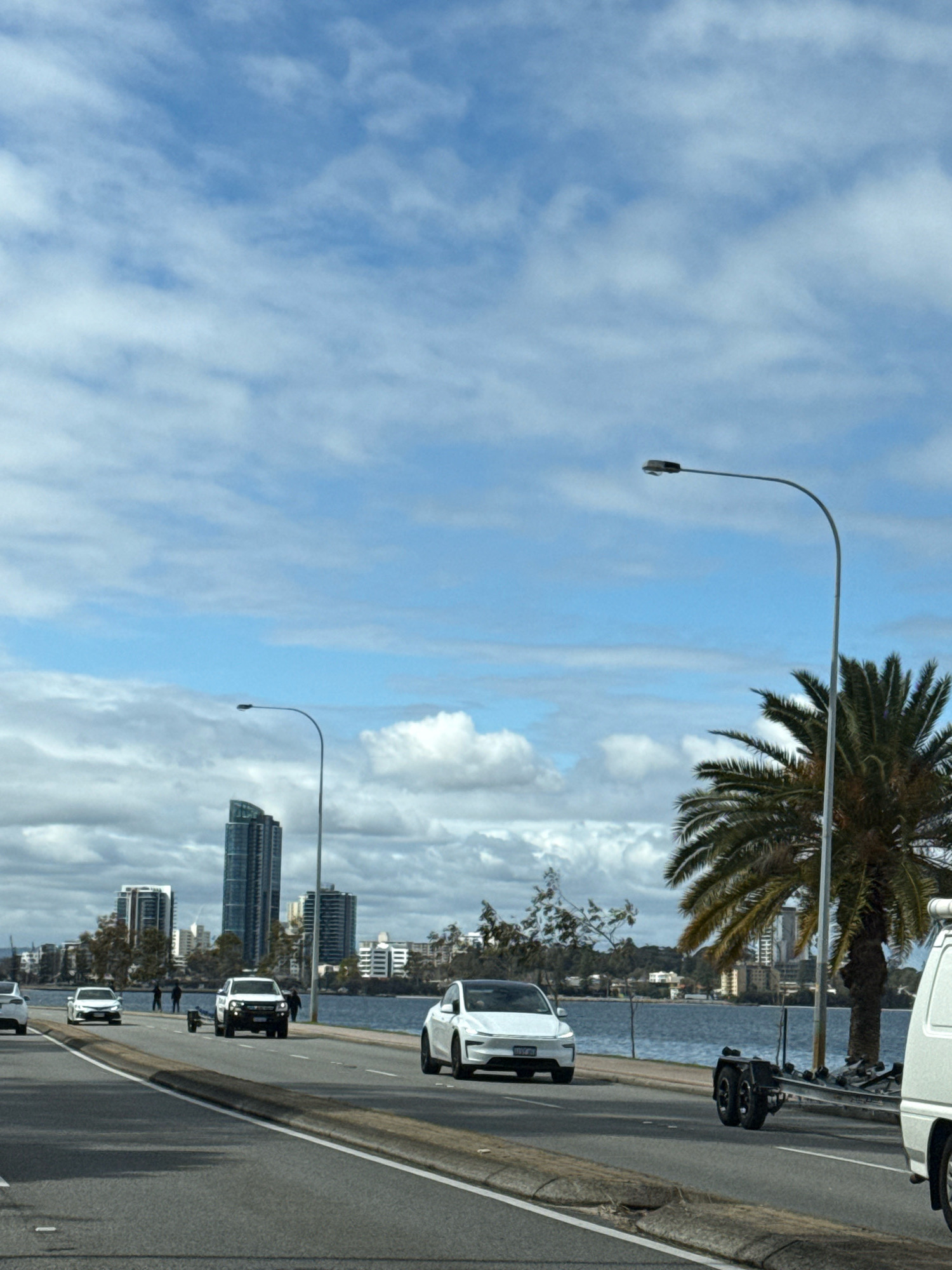The image shows a road in an urban waterfront area. On the road, several vehicles are visible, including a white Tesla, a white pickup truck towing a trailer, and other cars. In the background, there is a body of water with buildings and a cityscape on the other side. There are also palm trees and streetlights. The sky is partly cloudy.