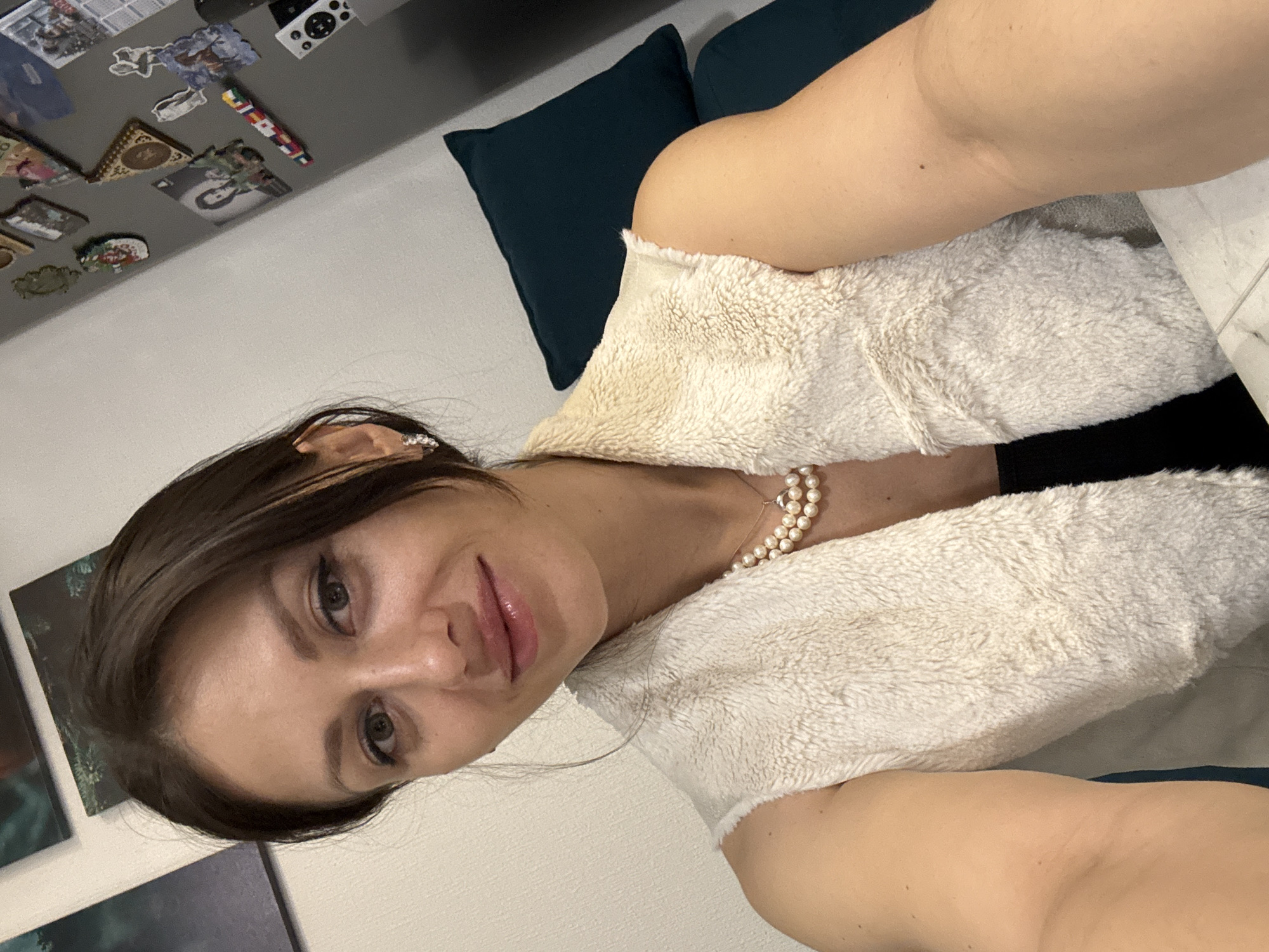 A woman is posing for a selfie. She is wearing a light-colored, furry vest over a black top and a pearl necklace. She has brown hair and is wearing earrings. She appears to be indoors, with a wall behind her decorated with various magnets. There are also dark blue cushions visible in the background. The location is potentially in Murino, Russia.